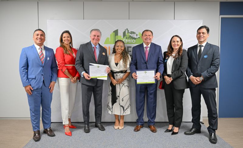 Foto horizontal: TJMA recebe menções honrosas do Prêmio Solo Seguro. A foto mostra um grupo de seis pessoas em pé, posando para uma foto em um evento formal. Eles estão alinhados na frente de um banner cinza com o nome "PRÊMIO SOLO SEGURO". O grupo é formado por quatro homens e duas mulheres, todos bem-vestidos com roupas formais. O homem à esquerda veste um terno azul claro. A mulher ao lado dele veste um blazer vermelho e calças brancas. Um dos homens no centro, de terno cinza, e o homem ao lado dele, de terno azul marinho, seguram prêmios ou certificados. A mulher no centro veste um vestido branco e preto. A mulher mais à direita veste um terno preto com uma camisa branca. Ao fundo, há um painel branco com o logo do prêmio e uma parede azul-clara. O ambiente parece ser uma sala de eventos.