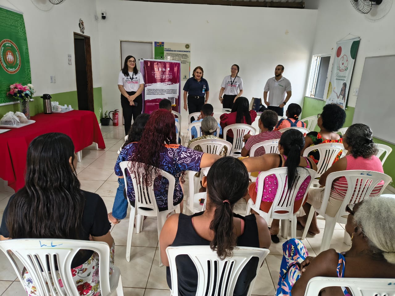 Sala clara, com paredes brancas e detalhes verdes, onde várias pessoas, majoritariamente mulheres, estão sentadas em cadeiras plásticas brancas, voltadas para a frente. Na frente da sala, quatro pessoas (três mulheres e um homem) estão em pé, conduzindo uma atividade. À esquerda, há uma mesa coberta por toalha vermelha com garrafa térmica, copos e arranjo de flores. Um banner do programa ¿Maria da Penha no Cotidiano¿ está exposto.