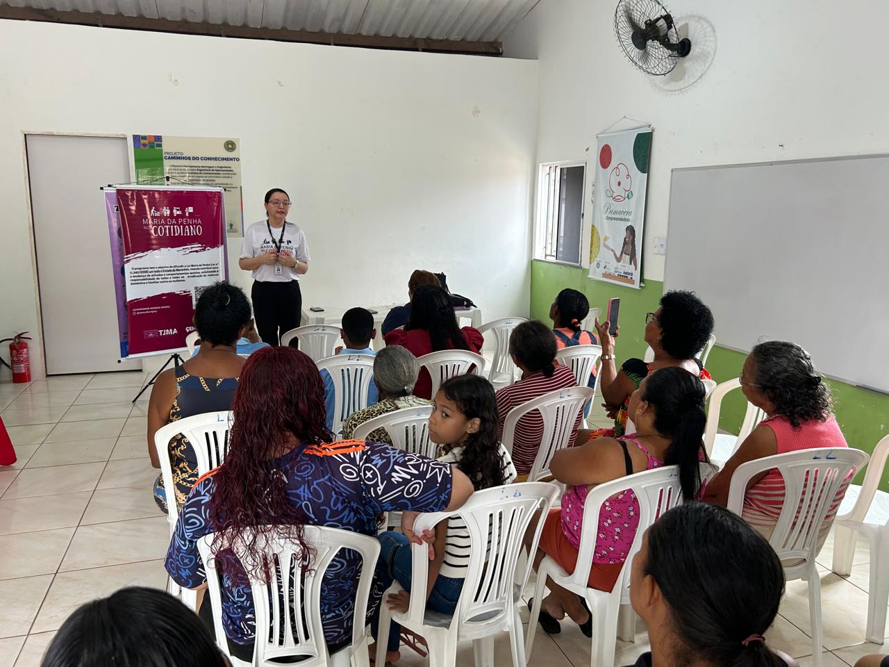 A mesma sala, com cadeiras plásticas brancas ocupadas por participantes. Uma mulher de óculos, camiseta branca do programa e crachá está em pé, falando para o grupo. À sua frente, um banner do ¿Maria da Penha no Cotidiano¿. Ao fundo, uma parede branca e uma janela aberta deixam entrar luz natural.