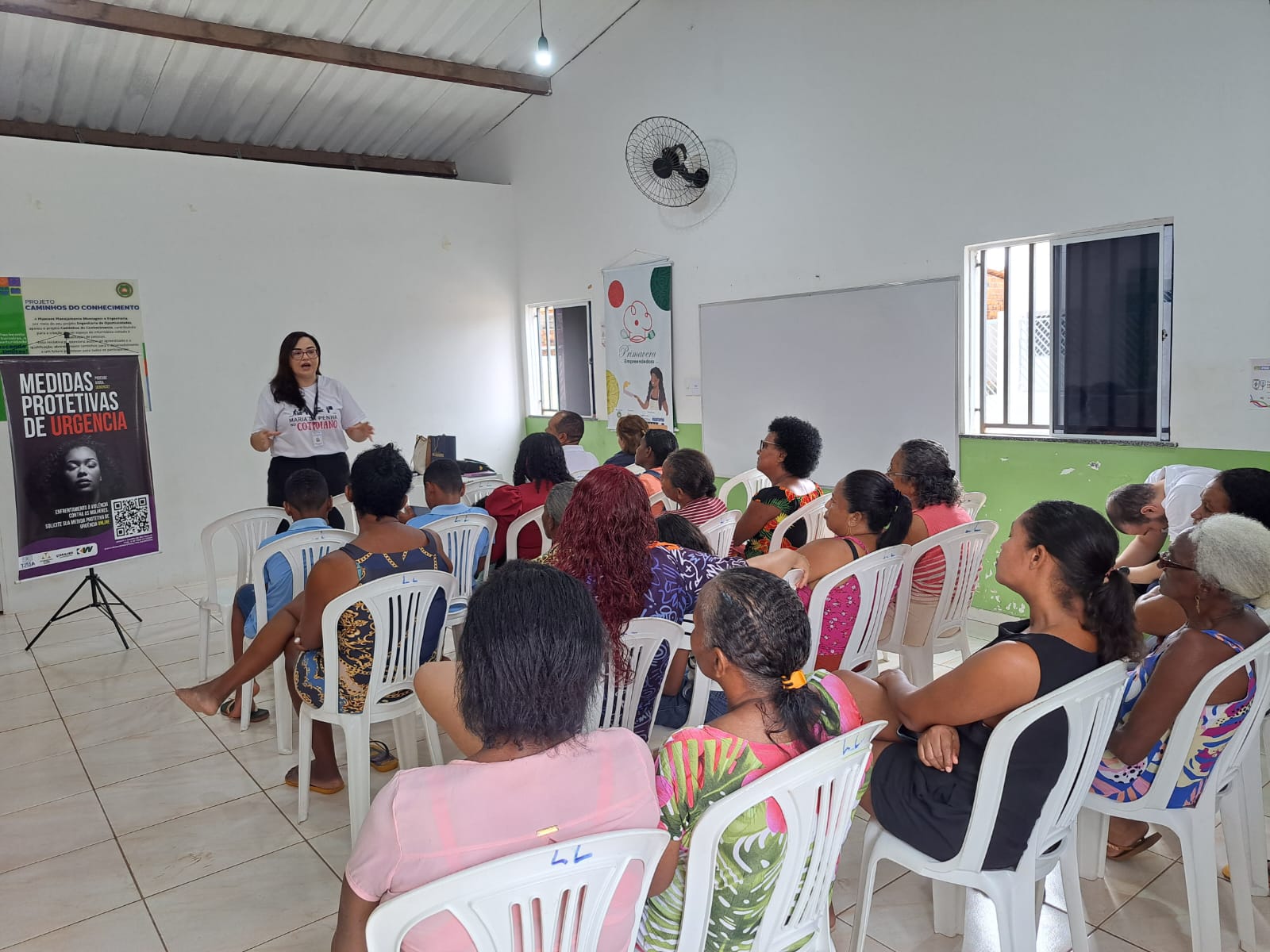 Sala semelhante às anteriores, com participantes sentadas em cadeiras plásticas brancas, voltadas para a frente. Uma mulher de cabelos longos e óculos, vestindo camiseta branca com a logo do programa e crachá, fala ao grupo. Ao lado dela, um banner preto com os dizeres ¿Medidas protetivas de urgência¿ e a imagem de uma mulher em preto e branco.