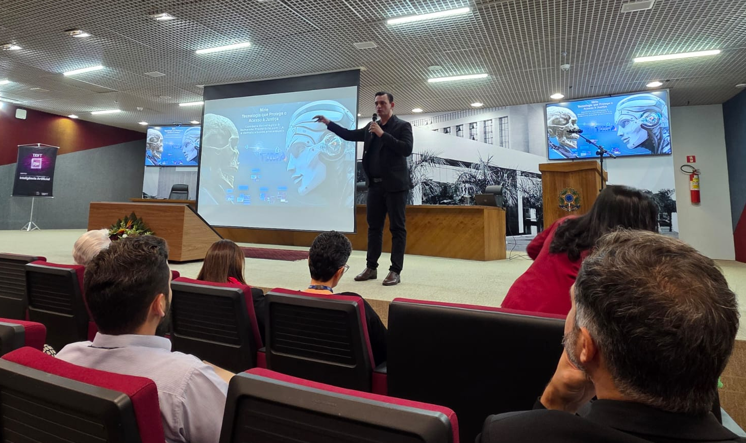 A imagem, capturada em um auditório, foca em um homem no palco, que gesticula com a mão direita enquanto segura um microfone com a esquerda. Ele está vestido com um terno preto e uma camisa preta. Atrás dele, uma grande tela de projeção exibe a apresentação "Nirie - Tecnologia que Protege o Acesso à Justiça" e o subtítulo "Combate Estratégico e Demanda Predatória com I.A. e demais abusos processuais". A apresentação exibe uma imagem que justapõe um crânio humano à esquerda com uma figura de robô à direita. Um logotipo da "TIMA" aparece no centro inferior da tela. Ao fundo, é possível ver parte da parede do auditório com um quadro em preto e branco.