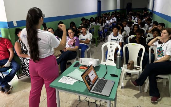 Foto horizontal de ações da Cemulher com campanha Agosto Lilás em Morros. A imagem mostra um ambiente de sala de aula, onde uma integrante da equipe está em pé na frente de um grupo de estudantes. A instrutora está de frente para os alunos, interagindo com eles. Os alunos estão sentados em fileiras, olhando para a frente, onde a representante da Cemulher está. Há uma mesa em primeiro plano com um notebook aberto.