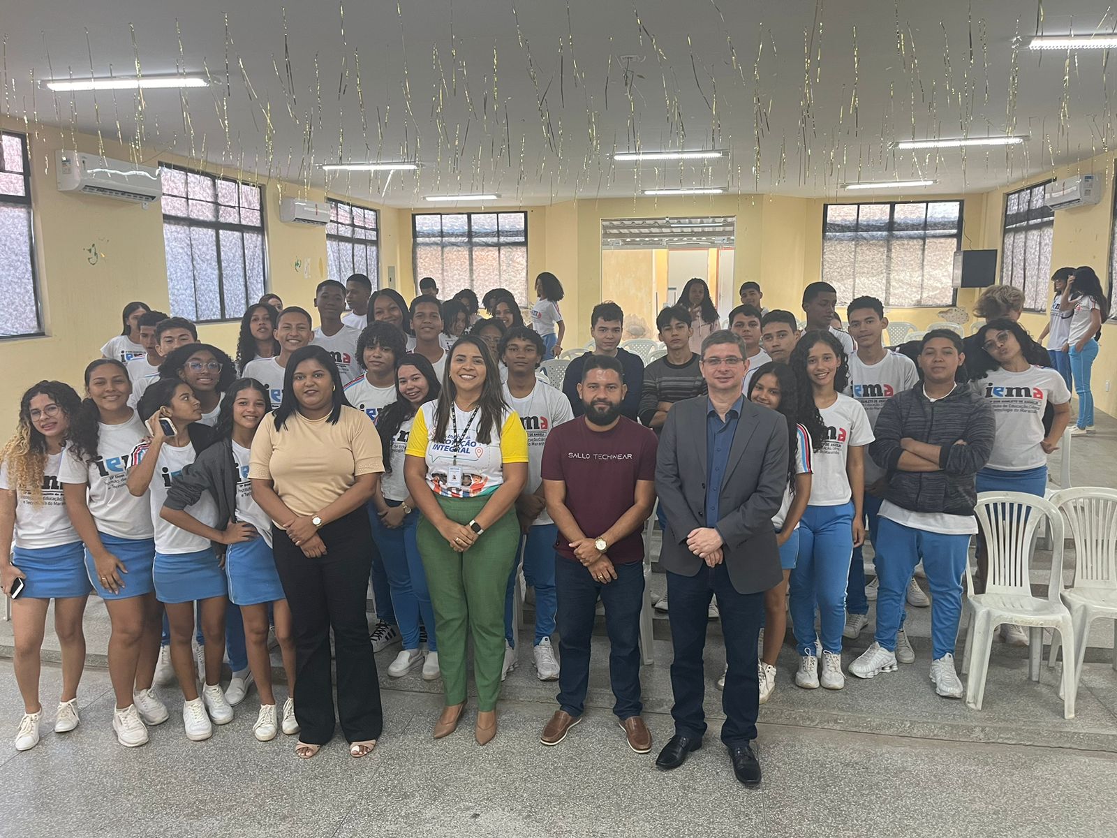 A imagem mostra um grupo de pessoas reunidas em um ambiente interno, provavelmente para uma foto em grupo. Os rostos estão cobertos por retângulos marrons para proteger a privacidade. A sala tem paredes brancas e está decorada com luzes de corda penduradas no teto. Há janelas com grades de metal e algumas cadeiras visíveis no primeiro plano.