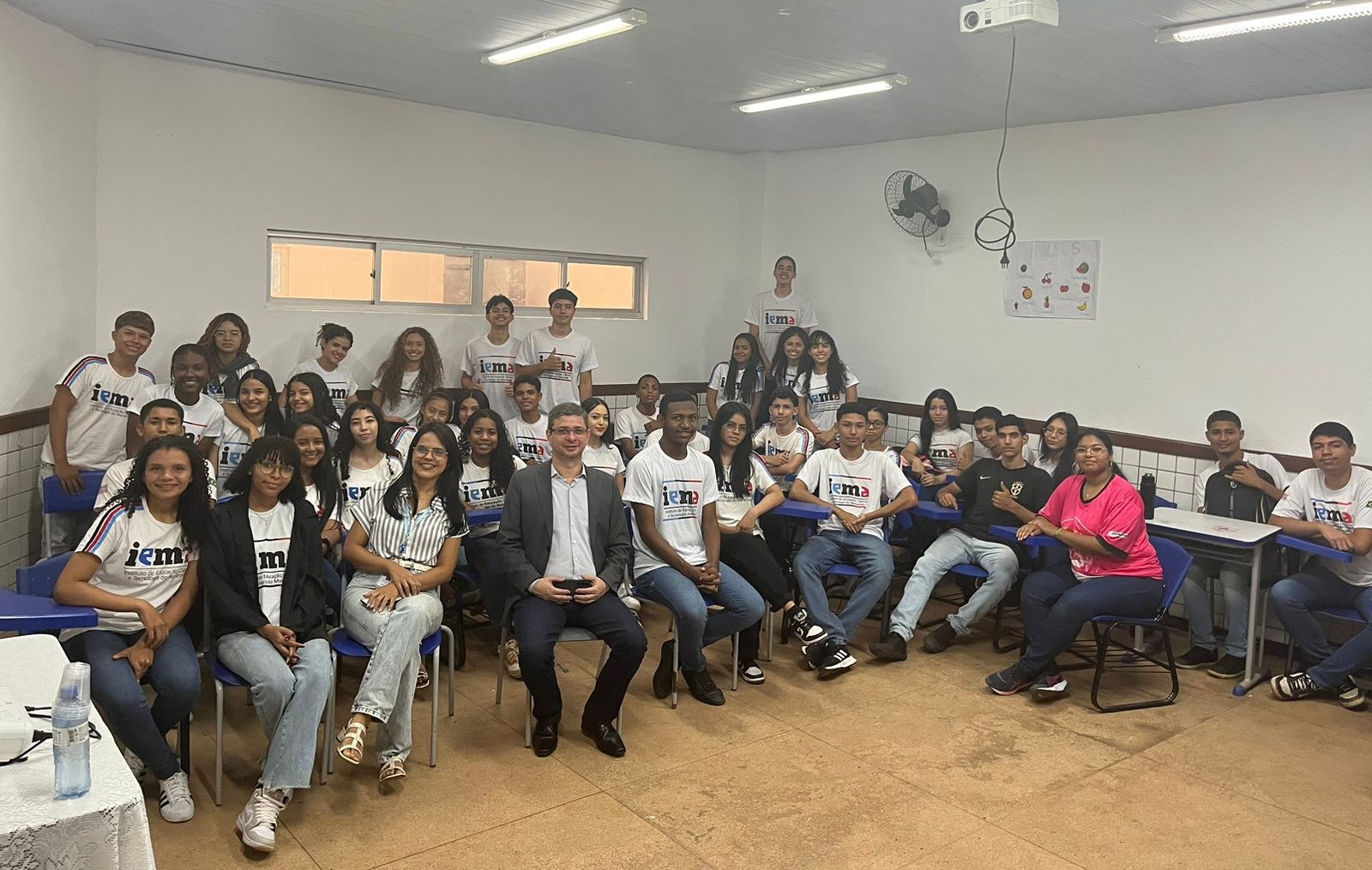 A imagem mostra um grupo de pessoas em uma sala de aula. Os rostos das pessoas estão obscurecidos com retângulos marrons para preservar a privacidade. Há mesas e cadeiras dispostas em fileiras, e as pessoas parecem estar vestindo camisas brancas semelhantes, com logotipos ou textos visíveis. Algumas pessoas estão em pé no fundo da sala, enquanto outras estão sentadas. Na parede, há um relógio e o que parece ser um papel ou pôster.