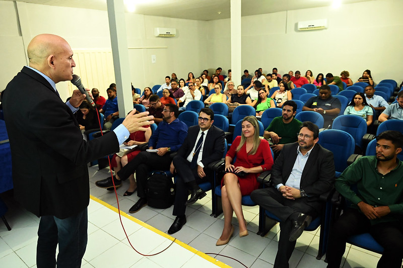 Foto horizontal do Curso de Regularização Fundiária em Timon, parte do programa Encontros Regionais. A imagem mostra o palestrante Daniel Pereira, homem branco de meia idade, de cabeça raspada, que usa blazer e calça jeans. Ele aparece à esquerda da foto, de perfil, falando ao público composto por homens e mulheres, sentados em cadeiras azuis, num auditório de paredes brancas, com duas coluna aparentes e dois aparelhos de ar condicionado.