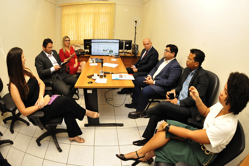 Foto horizontal de Encontros Regionais em Timon. Imagem mostra reunião de juízes com integrantes do Sistema de Justiça. Numa sala de Paredes amarelas e piso branco de cerâmica, com janela coberta por persiana ao fundo, homens e mulheres estão sentados em volta de uma mesa retangular de madeira marrom. Sobre uma das extremidades da mesa, há uma tela de computador. Todos os quatro homens usam terno, enquanto as três mulheres usam roupas variadas.