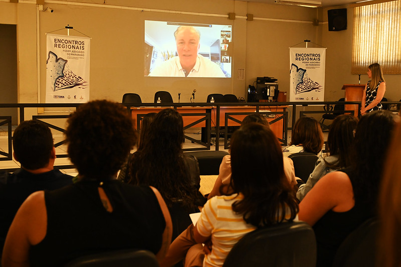 Foto horizontal da participação do presidente do TJMA, desembargador Froz Sobrinho no encerramento dos Encontros Regionais em Timon. A imagem mostra o Salão do Júri de Timon, com pessoas sentadas, assistindo a uma apresentação on-line do presidente numa tela projetada na parede.
A juíza Tereza Nina está à direita do telão, em pé, olhando em direção a ele. Nas laterais da sala, há banners com a inscrição ¿ENCONTROS REGIONAIS¿. As paredes são amarelas, há móveis na cor marrom e uma persiana em frente a uma janela, à direita.