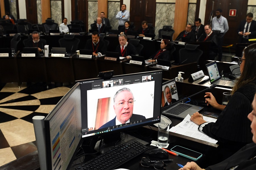 Foto horizontal de sessão do Órgão Especial em 4 de setembro de 2024. A imagem mostra a sala das sessões plenárias do TJMA. Há várias mesas com computadores e microfones, e pessoas sentadas em frente a elas. O foco da imagem está em um monitor de computador em primeiro plano, que exibe a imagem do presidente do TJMA, desembargador Froz Sobrinho.