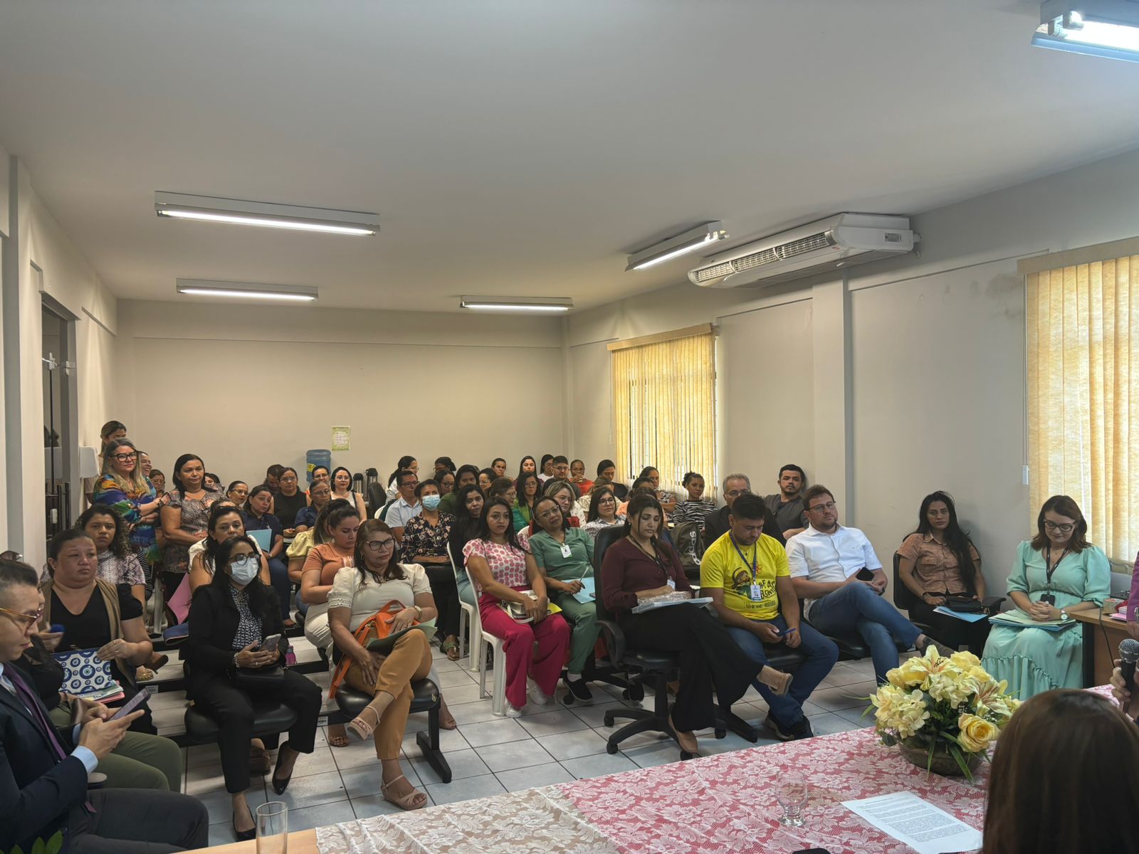 A imagem mostra uma reunião em um ambiente interno com várias pessoas sentadas e algumas em pé. As cadeiras estão organizadas em filas voltadas para a frente da sala, onde há uma mesa com um arranjo floral e uma toalha rosa. Uma pessoa está de pé na frente, aparentemente falando com o público. A sala tem paredes brancas, janelas com persianas parcialmente fechadas e iluminação fluorescente. Parece ser um evento ou reunião formal, dado o número de participantes e a organização do espaço.