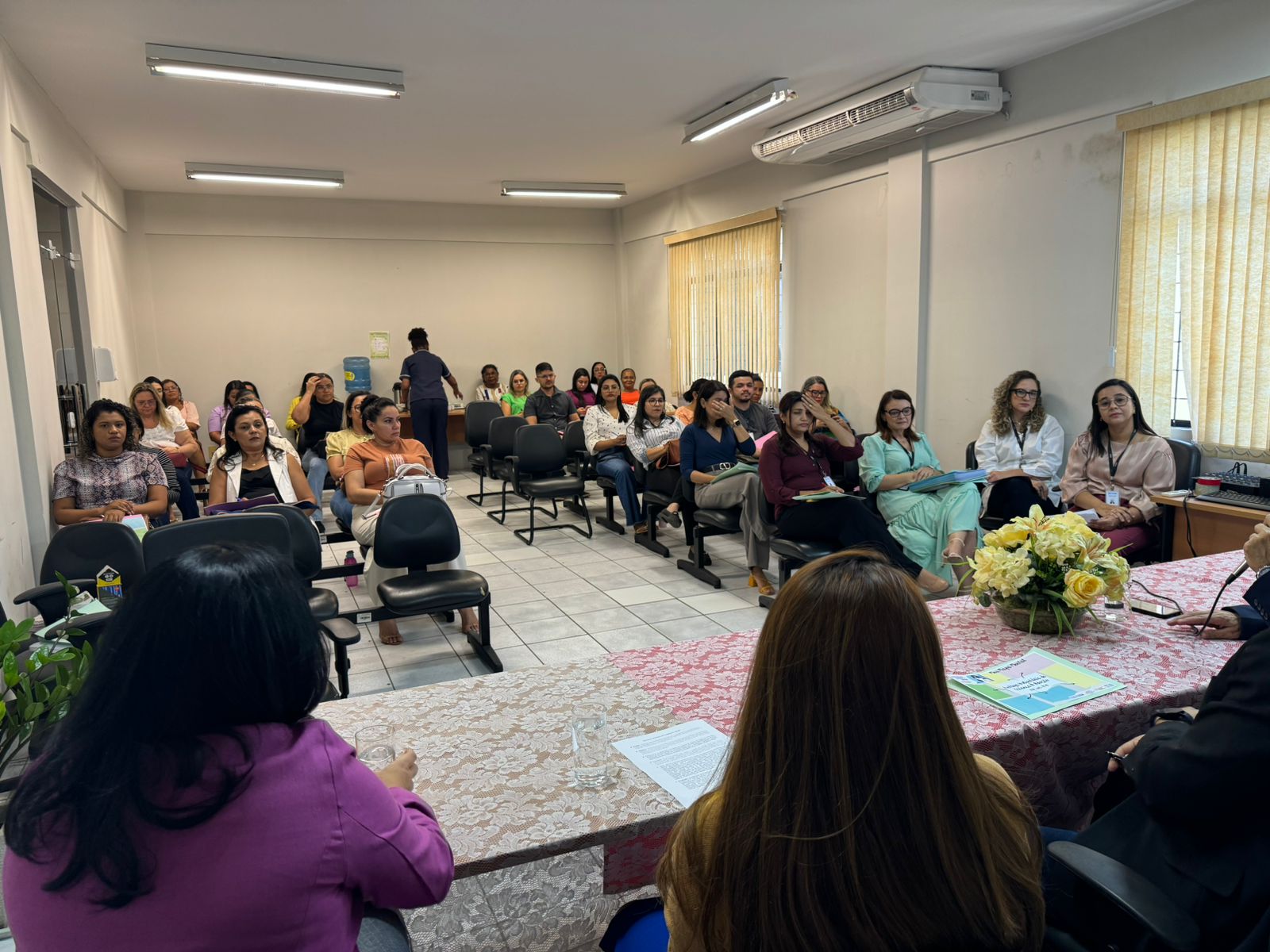 A imagem também mostra um grupo de pessoas sentadas em uma sala, aparentemente participando de uma reunião ou seminário. A sala tem paredes de cor creme e há uma mesa na frente com flores sobre ela. Os participantes estão sentados em cadeiras organizadas de frente para a mesa, onde parece haver uma apresentação ou palestrante, embora o foco não esteja em nenhum indivíduo específico. Algumas pessoas estão segurando papéis ou cadernos.