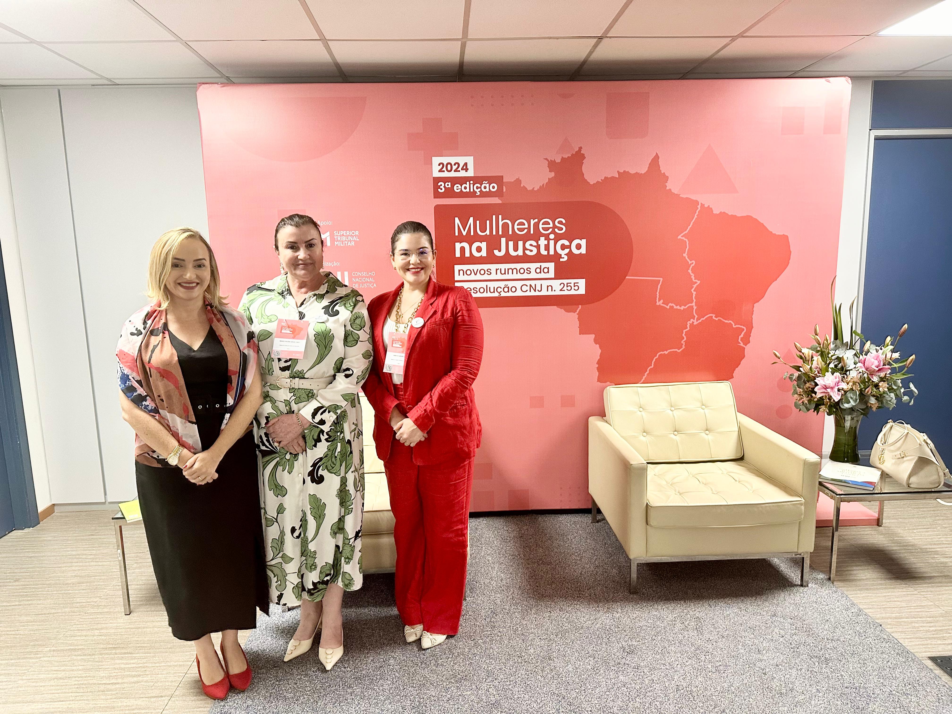 A imagem mostra três pessoas em pé em frente a uma parede rosa com textos e gráficos. A parede apresenta o contorno de um mapa do Brasil, alguns textos em português e o número ¿2024¿. As pessoas estão posicionadas à esquerda da imagem, cobrindo parte do conteúdo da parede. Elas parecem estar conversando ou posando para a foto. À direita da imagem, há um sofá branco.