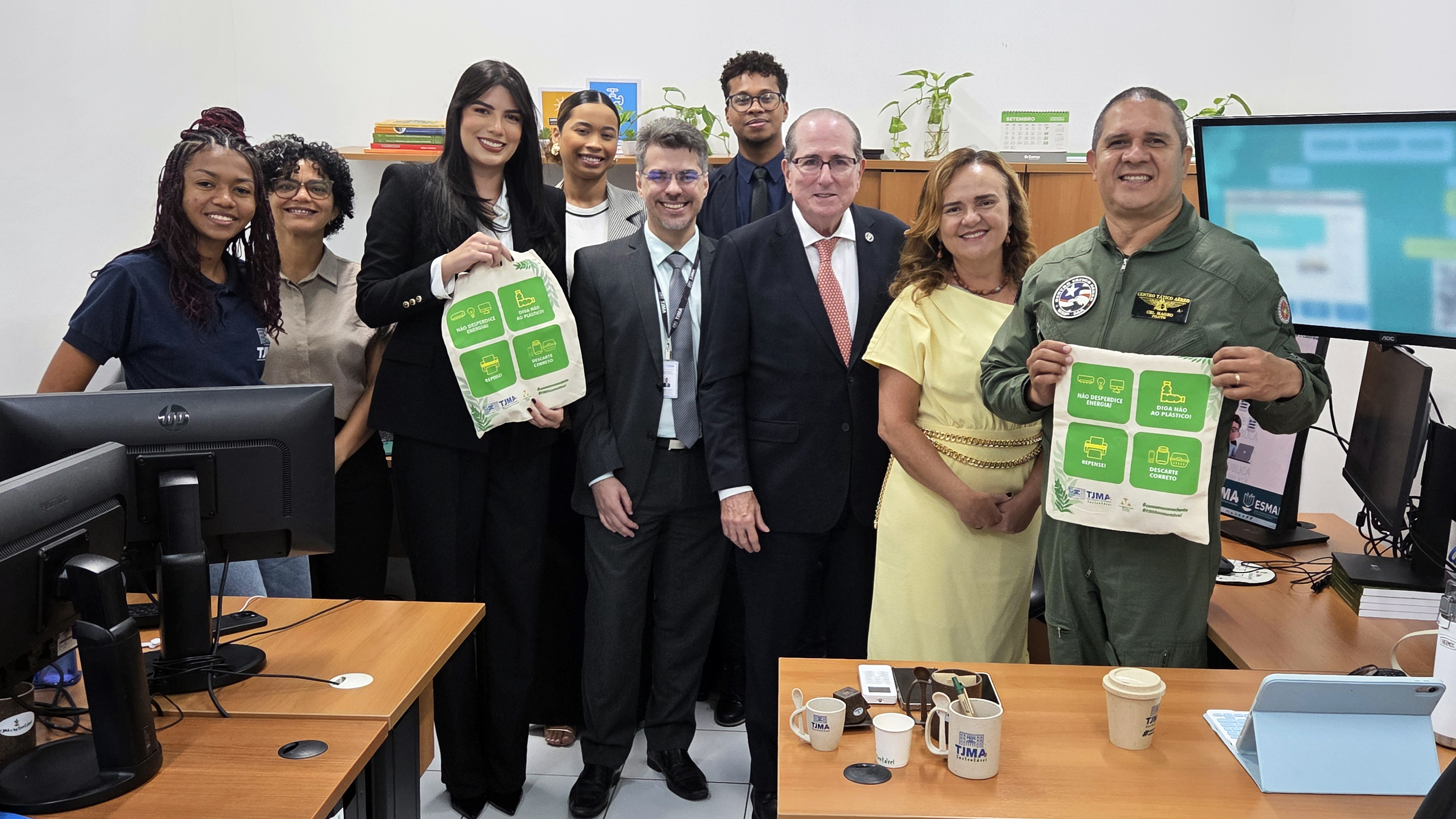 A imagem mostra um grupo de dez pessoas reunidas em um ambiente de escritório. Eles estão em pé, posando para a foto com expressões amigáveis e descontraídas. O espaço contém mesas com computadores, teclados e objetos de uso cotidiano, como copos e papéis. Dois participantes, à direita na frente, seguram ecobags com estampas verdes e brancas que remetem à sustentabilidade ¿ com ícones e textos relacionados à conscientização ambiental. Um deles veste um macacão verde com distintivos, indicando vínculo com aviação ou forças de segurança, provavelmente do Centro Tático Aéreo (CTA). Os demais estão em trajes sociais ou casuais, sugerindo uma reunião institucional. Ao fundo, há plantas sobre uma estante, reforçando o clima de cuidado ambiental. A imagem transmite um momento de integração entre diferentes setores, possivelmente celebrando ou promovendo uma iniciativa conjunta voltada à sustentabilidade.