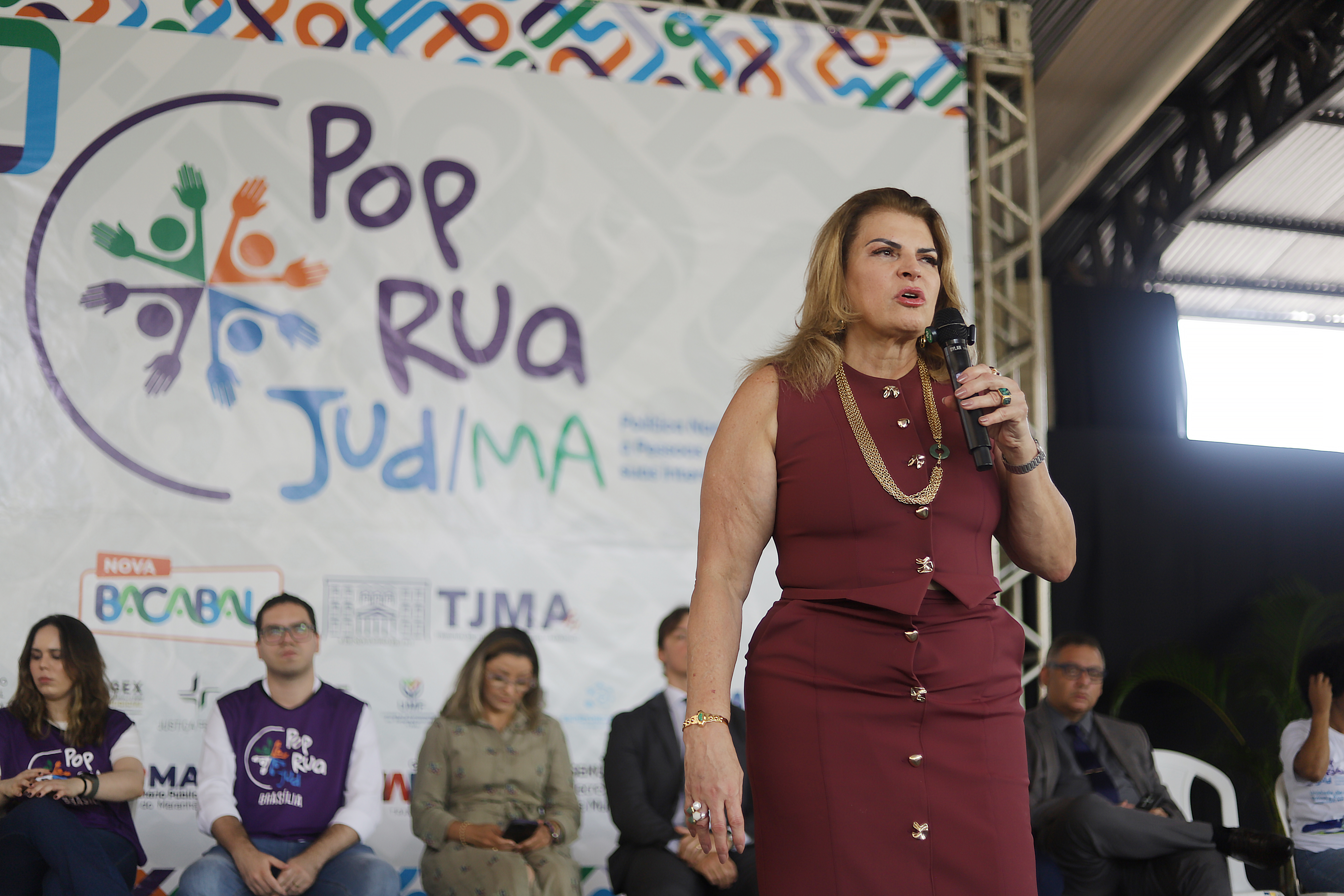 Mulher fala ao microfone em um palco, vestindo um conjunto vinho. Atrás dela há um painel com o logo do projeto PopRuaJud/MA. Outras pessoas estão sentadas no palco, observando a fala.