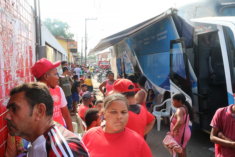 Foto horizontal do evento PopRuaJud em São João Batista.  Há um grande número de pessoas de diferentes idades na rua, muitas delas vestindo camisas de cores vibrantes, principalmente vermelho. A cena se passa em uma rua entre uma parede de tijolos e o ônibus de atendimento. O muro à esquerda é de cor clara com detalhes em vermelho, e a rua tem trânsito de motos ao fundo. Um ônibus grande e adaptado está estacionado à direita, com a porta aberta e o toldo estendido. A lateral do veículo é predominantemente azul.