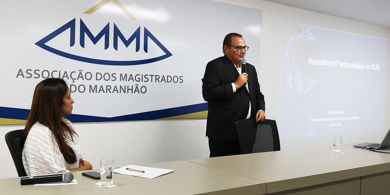 Foto colorida horizontal do palestrante, o gestor Sandro Monteiro, sentado, falando ao microfone, no auditório da AMMA durante o Seminário de Saúde Pública e Suplementar. Ao lado dele está a mediadora Mirela Rego.