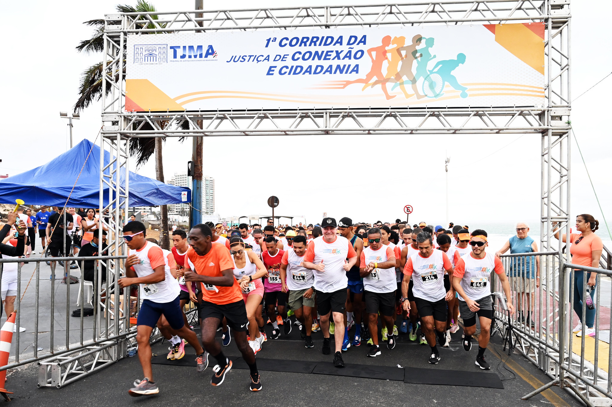 A imagem mostra um grupo de corredores na linha de partida de uma corrida, prestes a começar. Eles estão posicionados sob uma grande faixa que diz ¿1ª CORRIDA DA JUSTIÇA DE CONEXÃO E CIDADANIA¿. A cena é ao ar livre, com barricadas ao longo dos lados da área de partida. Os corredores estão usando várias roupas atléticas adequadas para corrida, e alguns têm números presos às suas roupas, indicando sua participação na corrida. Esta imagem captura o momento antes do início de um evento esportivo, mostrando um evento comunitário ou competitivo organizado, focado em fitness e possivelmente na conscientização ou arrecadação de fundos para justiça, conexão e cidadania, como sugerido pelo texto da faixa.