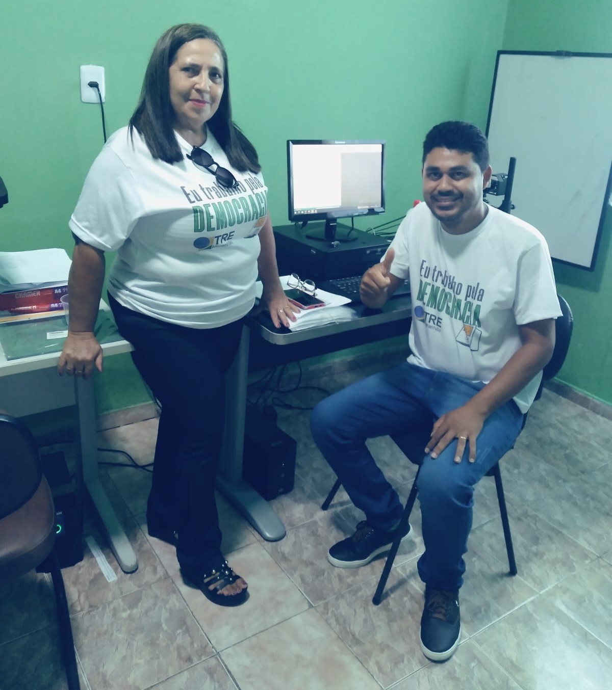 Foto da Sala do Justiça de Todos 4, durante as eleições 2024. A imagem mostra duas pessoas na sala: uma mulher e um homem. A pessoa à esquerda está em pé, vestindo uma camiseta branca com texto, calças escuras e sandálias. A pessoa à direita está sentada, também vestindo uma camiseta branca com texto, jeans e tênis. Ao fundo, há uma tela de computador em uma mesa e uma tela numa parede verde.