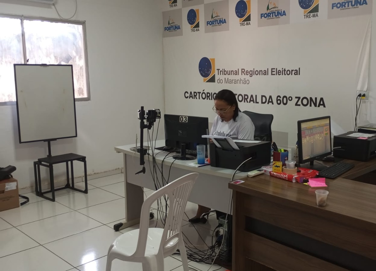 Foto da Sala do Justiça de Todos em Fortuna durante as eleições 2024. A imagem mostra sala, que funcionou como Cartório Eleitoral da 60ª Zona do Tribunal Regional Eleitoral do Maranhão. Há uma mesa com um computador, monitor, teclado, mouse e vários materiais, como canetas e grampeadores. No fundo, há um quadro branco ao lado de uma janela com persianas fechadas. Também há cadeiras em frente à mesa. Há uma mulher de meia idade, usando óculos de grau, sentada em frente ao monitor.