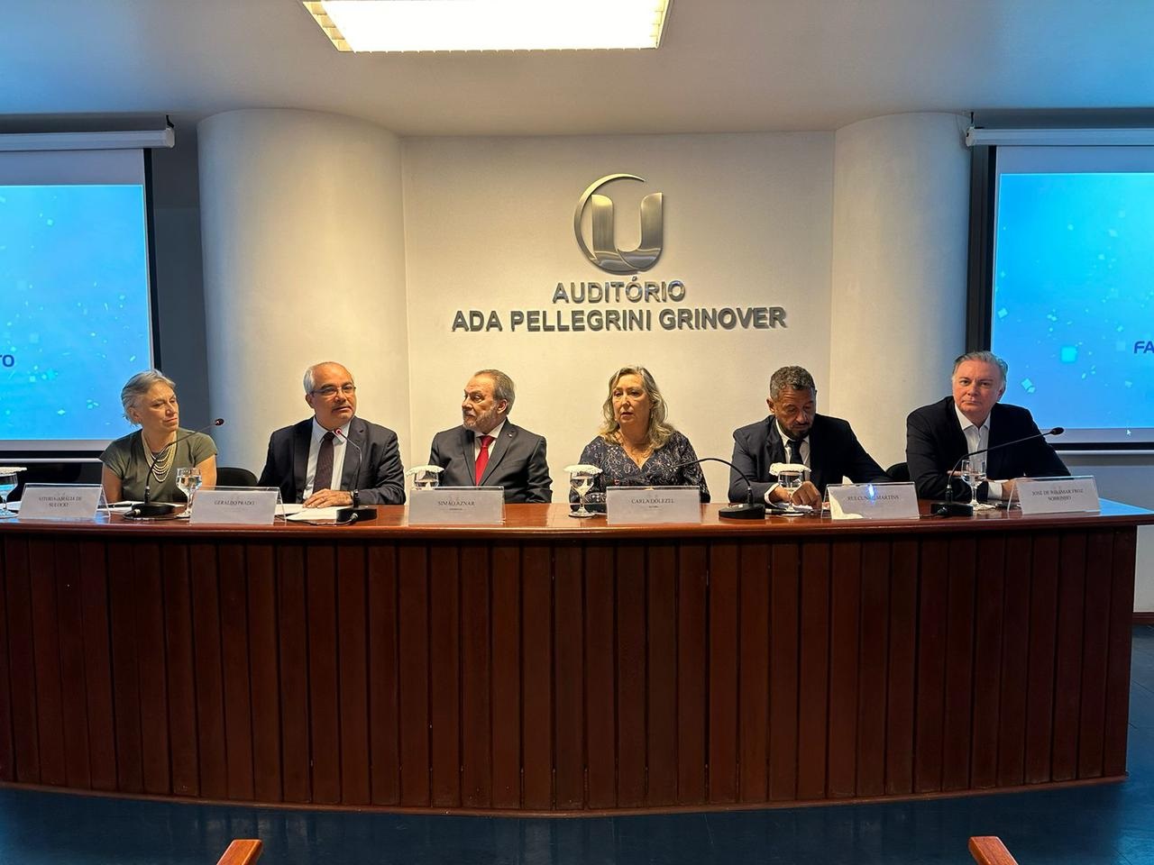 Foto horizontal de participação do TJMA em evento sobre processo penal e contemporaneidade. A imagem mostra um grupo de pessoas sentadas em uma longa mesa durante a conferência. O presidente do TJMA, desembargador Froz Sobrinho, está na extremidade direita da foto, esquerda da mesa. Atrás das pessoas, há uma parede com o nome "AUDITÓRIO ADA PELLEGRINI GRINOVER", indicando o nome do auditório onde o evento está ocorrendo. Há microfones na frente de cada pessoa. A sala tem grandes painéis.