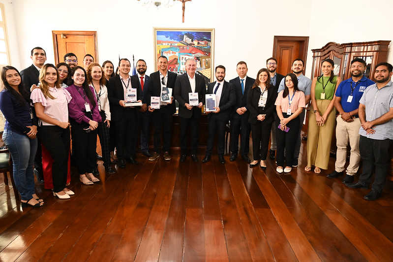 Foto horizontal da entrega do prêmio Prática 5 estrelas. A imagem mostra um grupo de pessoas posando para uma foto em uma sala. A sala tem piso de madeira e duas portas de madeira ao fundo. No centro da parede do fundo, há uma pintura colorida de uma paisagem urbana. Acima da pintura, há um crucifixo. As pessoas, com o presidente do TJMA, desembargador Froz Sobrinho ao centro, estão organizadas em duas fileiras, com algumas segurando documentos e prêmios. Algumas das pessoas estão usando crachás de identificação. A sala também contém um móvel de madeira à direita.