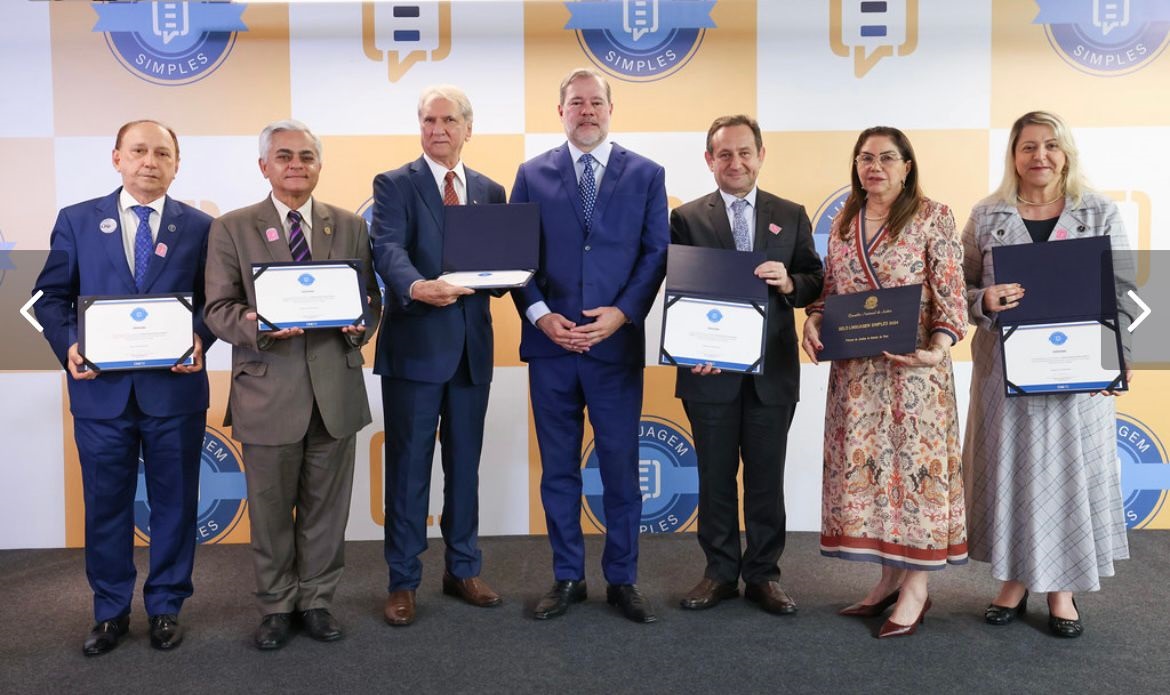 Foto horizontal da cerimônia de entrega do Selo Linguagem Simples, com presença do desembargador Lourival Serejo e representantes de outros tribunais premiados. A imagem mostra sete pessoas em pé, lado a lado, segurando os certificados do prêmio. Eles estão em um ambiente formal na sede do STF. O fundo é composto por um padrão quadriculado em tons de amarelo e branco, com logotipos azuis que contêm a palavra "SIMPLES". As pessoas estão vestidas formalmente, com ternos e vestidos.