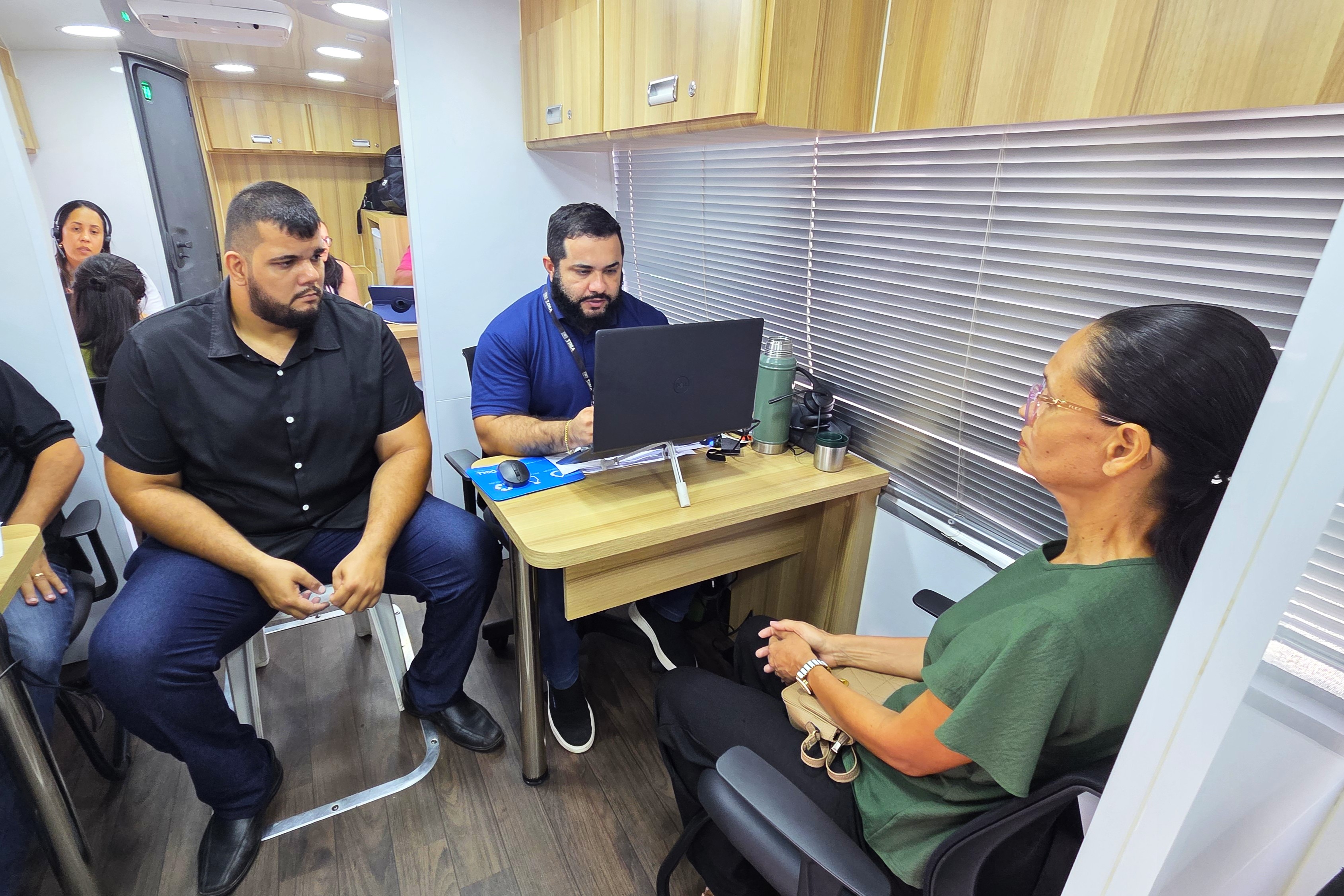 ## ¿ Descrição da Imagem  A imagem captura um momento de **atendimento individualizado** no interior do ônibus de cidadania, o mesmo ambiente visto na foto anterior.  **Cena Principal (Atendimento):**  * **Mulher em Atendimento (Direita):** Uma mulher, provavelmente a cidadã sendo atendida, está sentada em uma cadeira preta, de perfil para a câmera. Veste uma camisa polo verde escura e óculos de grau. Ela tem o cabelo preso e está olhando e gesticulando em direção aos atendentes, com as mãos cruzadas no colo. * **Atendente (Centro):** Um homem de barba, usando uma camisa polo escura (azul marinho ou preta) com uma etiqueta ou crachá, está sentado em frente a um pequeno balcão/mesa. Ele está focado em um laptop e interagindo com a mulher, indicando que está registrando informações ou prestando o serviço solicitado. * **Segundo Atendente/Observador (Esquerda):** Um homem corpulento, de camisa social preta de mangas curtas e calças jeans escuras, está sentado ao lado do balcão, observando o atendimento. Ele segura algo pequeno nas mãos (possivelmente um objeto pessoal ou um pedaço de papel). Sua presença sugere que ele pode ser um segundo funcionário ou um supervisor/observador do processo.  **Ambiente:**  * **Interior:** O ambiente é o mesmo do ônibus adaptado, com piso de madeira escura e armários superiores de madeira clara. * **Detalhes da Estação de Trabalho:** A mesa tem um laptop em um suporte elevado. Há uma garrafa térmica escura e um mousepad azul. * **Janela/Persiana:** Uma grande persiana horizontal de cor cinza-clara cobre a janela lateral do veículo, controlando a luz externa. * **Fundo:** Uma mulher pode ser vista no canto superior esquerdo, possivelmente aguardando ou passando por outro posto de atendimento, adicionando profundidade à cena.  **Em resumo, a foto ilustra a fase prática da ação de cidadania, mostrando o trabalho dos servidores em prestar um serviço ou informação a uma cidadã dentro da estrutura móvel do ônibus.**  ---  Gostaria de procurar por notícias ou mais detalhes sobre a ação do TJMA para entender o **tipo de serviço** que está sendo oferecido neste momento?