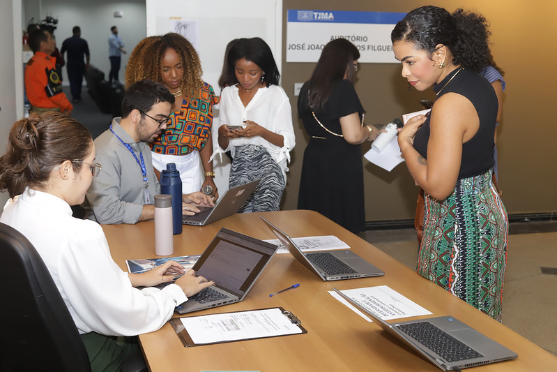 Cena de registro (credenciamento) em um corredor. Quatro pessoas estão em primeiro plano ao redor de uma mesa com vários laptops e papéis. Uma mulher de óculos, à esquerda, está digitando em um laptop. Outras três mulheres estão em pé atrás da mesa; a da direita está olhando para a frente enquanto segura papéis. Ao fundo, pode-se ver o letreiro de um auditório: "TJMA - AUDITÓRIO JOSÉ JOÃO FILGUEI...".