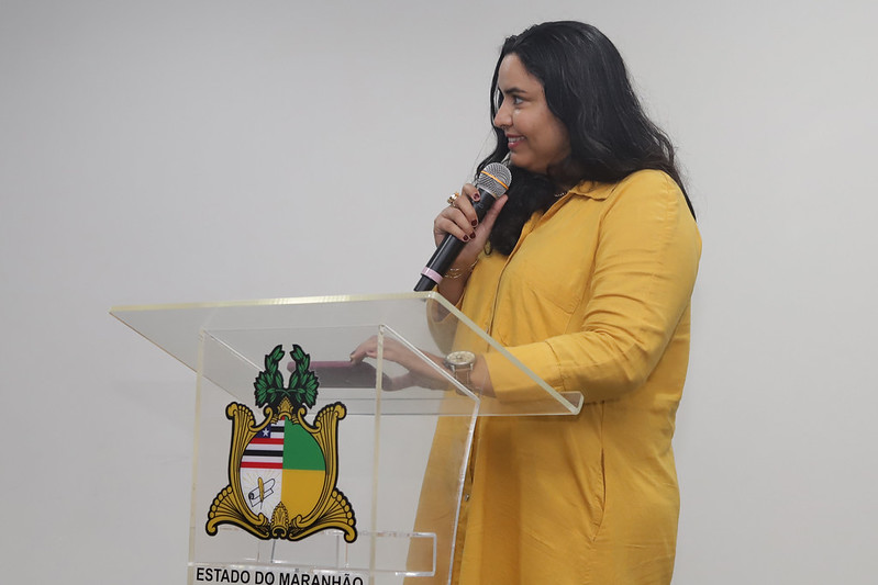 Uma mulher jovem, de cabelos escuros e longos, vestindo um vestido-camisa amarelo mostarda, está sorrindo enquanto fala em um microfone que segura com a mão direita. Ela está de pé ao lado do mesmo púlpito de acrílico com o brasão do Maranhão. O fundo é uma parede branca.