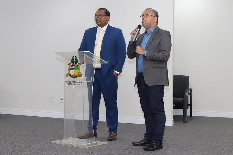 Dois homens estão em pé ao lado do púlpito de acrílico. O homem à esquerda veste um terno azul-marinho com gravata amarela clara. O homem à direita, com barba, veste um blazer cinza-chumbo sobre uma camisa azul e segura um microfone com a mão esquerda, falando.