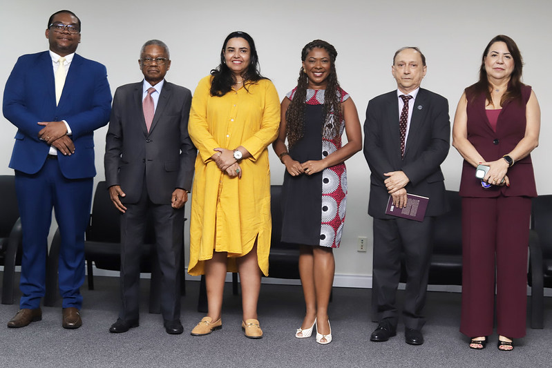 Seis pessoas, três homens e três mulheres, estão alinhadas lado a lado em um palco para uma foto de grupo. Todos estão em trajes sociais ou executivos. Da esquerda para a direita: um homem de terno azul, um homem mais velho de terno cinza, a mulher de vestido amarelo (vista anteriormente no púlpito), uma mulher de vestido preto e branco estampado, o homem de terno cinza-escuro (visto anteriormente no púlpito), e uma mulher de terno vinho-escuro.