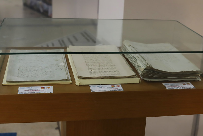 Vista em close-up de uma vitrine de vidro contendo documentos históricos ou antigos. Dentro da vitrine, há três conjuntos de papéis amarelados e manuscritos. À esquerda, um conjunto menor de papéis abertos está acompanhado de uma placa de identificação que menciona "TJMA" e o título "TOMADA DE VASC... MARANHÃO". No centro, há outro conjunto de papéis abertos com uma placa de identificação que menciona "TJMA" e a descrição incompleta "JUIZ DO ENCARREGADO MUNICIP...". À direita, há uma pilha espessa de documentos empilhados, também com uma placa do "TJMA" e uma descrição incompleta. A vitrine está sobre uma base de madeira clara.