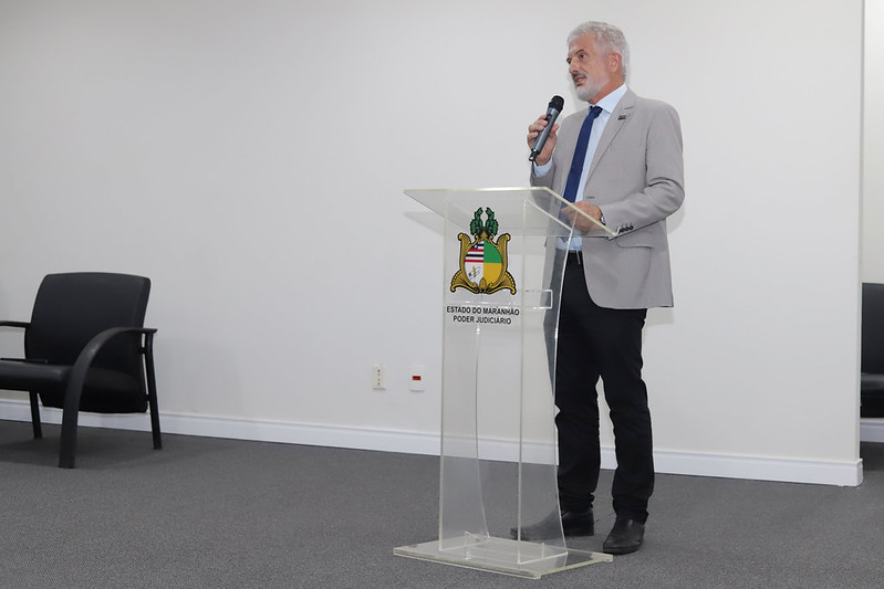 Um homem de cabelos grisalhos e barba bem-cuidada, vestindo um blazer cinza-claro sobre uma camisa social azul-escura e gravata, está em pé atrás de um púlpito de acrílico transparente. Ele segura um microfone com a mão direita e está falando. No centro do púlpito, é visível o brasão do Estado do Maranhão - Poder Judiciário. O fundo é uma parede branca lisa e o piso é um carpete cinza. Uma cadeira de escritório preta com braços curvos é vista no lado esquerdo da imagem, em um plano mais próximo.
