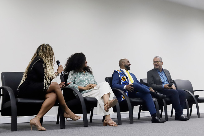 Quatro pessoas sentadas em uma fileira de cadeiras pretas em um palco, participando de um painel de discussão. Uma mulher de costas para a câmera, vestida de preto, segura um microfone e fala com as outras. As outras três pessoas (uma mulher de blusa estampada, um homem de terno azul e um homem de blazer cinza) estão sentadas e engajadas na conversa.