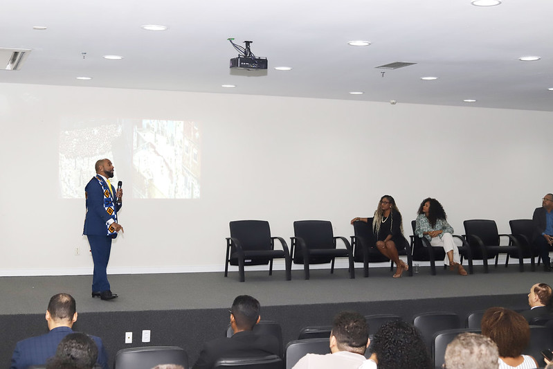 Uma cena de painel em um auditório. Um homem de terno azul-marinho com uma estola colorida de padrão geométrico e gravata amarela está em pé no palco, falando, com uma imagem projetada na parede branca atrás dele. À direita, três mulheres estão sentadas em cadeiras pretas esperando sua vez de falar. A plateia está visível em primeiro plano, com pessoas sentadas assistindo.