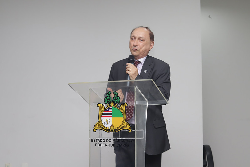 Um homem de meia-idade, com cabelos castanhos curtos e usando um terno cinza-escuro com gravata vinho estampada, está em pé atrás de um púlpito de acrílico transparente. Ele segura um microfone com a mão direita e gesticula levemente com a esquerda. No centro do púlpito, está o brasão do Estado do Maranhão - Poder Judiciário. O fundo é uma parede branca simples.