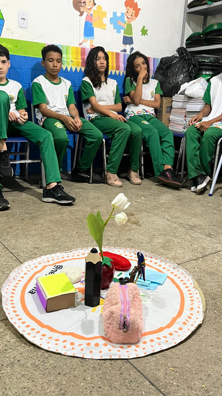 grupo de adolescentes usando uniforme escolar em cores verde e branco, sentados próximos a um circulo feito de objetos no chao