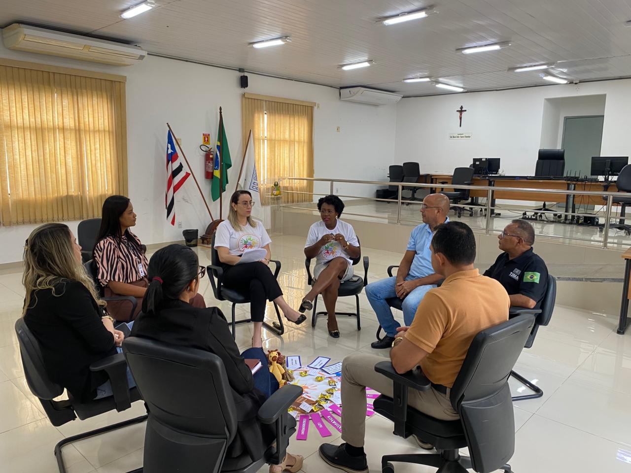 em ambiente interno, um grupo de pessoas entre homens e mulheres sentados e dispostos em formato circular, participam de atividade com objetos dispostos no chao no centro da ação