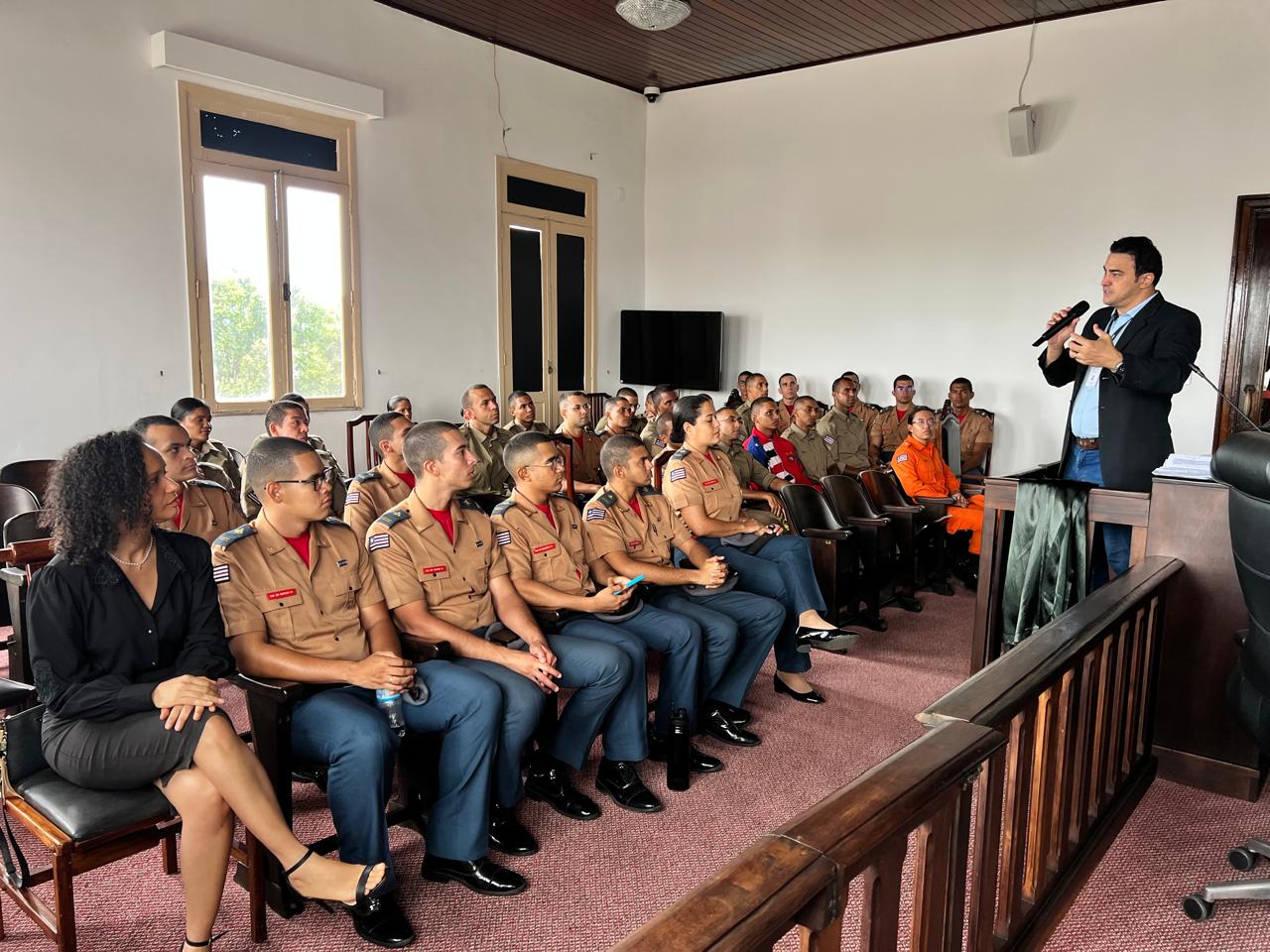 Sala ampla e iluminada, com paredes brancas e janelas altas. Diversos cadetes do CFO, uniformizados em tons de bege e azul, estão sentados em fileiras, atentos. À direita da imagem, um servidor do TJMA fala ao microfone, de pé, voltado ao grupo. À esquerda, uma mulher de roupa preta observa a apresentação.