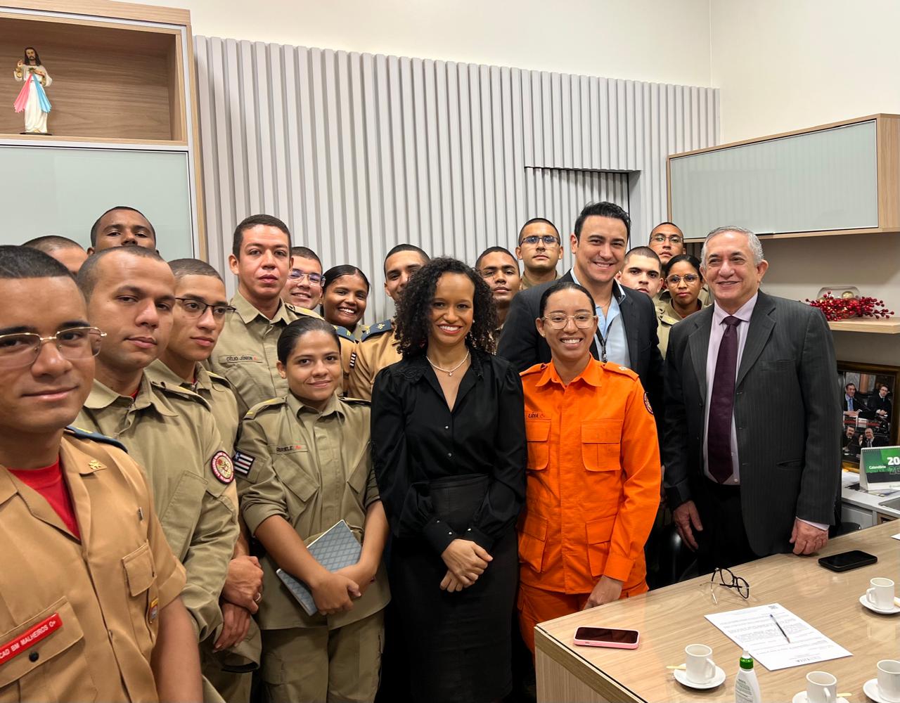 Ambiente de gabinete institucional, com mesa de madeira à frente e quadros na parede. Um grupo de cadetes uniformizados ocupa quase toda a imagem, posando para foto. No centro, duas pessoas ¿ uma mulher de roupa preta e uma cadete de uniforme laranja ¿ aparecem sorrindo. Um servidor do TJMA e um desembargador também estão presentes, à direita.