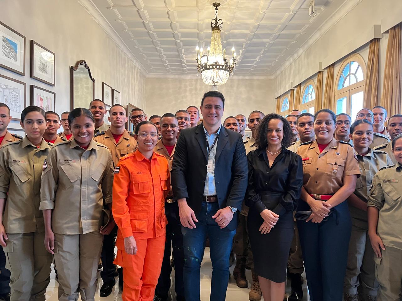 Salão nobre do TJMA, com lustre grande pendurado no teto e cortinas douradas ao fundo. Um grupo numeroso de cadetes uniformizados está posicionado para foto. No centro, estão uma cadete com uniforme laranja, um servidor do TJMA de paletó escuro e uma mulher de roupa preta. Todos posam sorrindo.