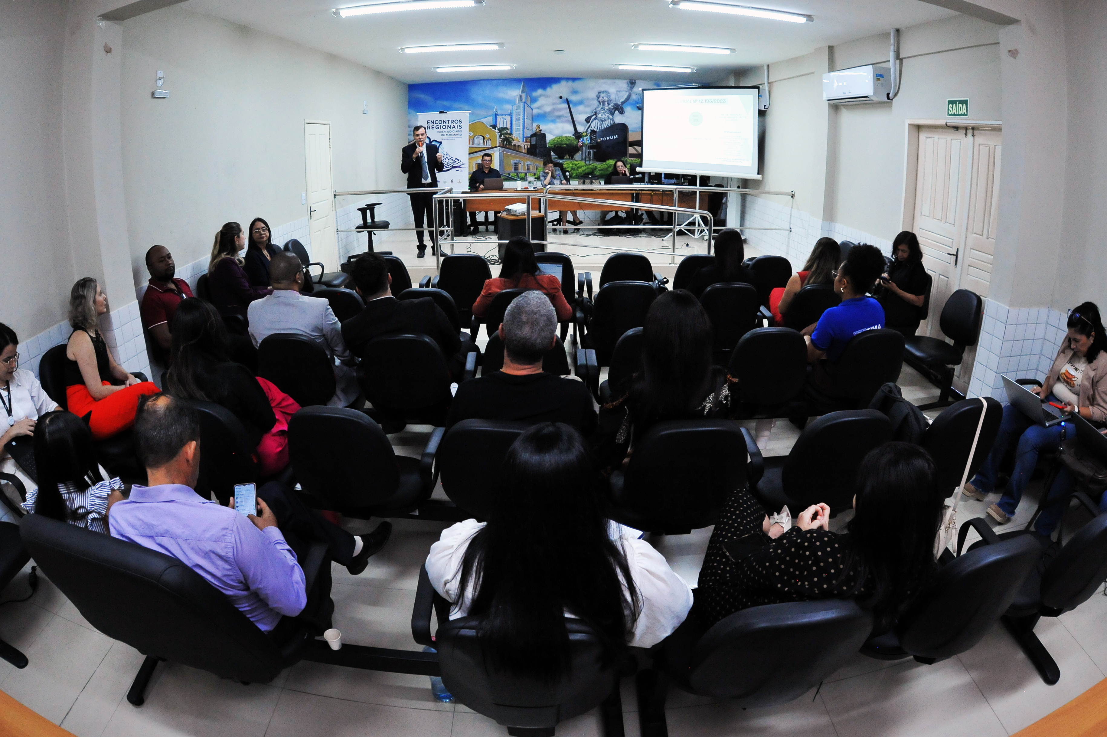 Esta imagem mostra uma cena de uma **apresentação ou evento em um ambiente fechado**, possivelmente um **auditório ou sala de reuniões**. Aqui estão os detalhes observados: * **Pessoa Principal/Palestrante:** Um homem de barba, vestindo um terno escuro (azul marinho ou preto) e camisa branca, está em pé no centro superior da imagem, **falando em um microfone** de mão. Ele parece ser o palestrante ou apresentador. * **Público:** Várias pessoas estão sentadas em cadeiras escuras, voltadas para o palestrante. A maioria das pessoas é vista de costas ou parcialmente, com foco nas nucas e ombros, sugerindo que estão atentas à apresentação. * **Cenário/Fundo:** Atrás do palestrante, há um **painel ou banner de fundo**. A parte superior deste painel tem um texto que diz "ENCONTROS REGIONAIS DO JUDICIÁRIO" (ou similar, dependendo da resolução). A maior parte do painel é uma **imagem colorida com elementos arquitetônicos** que parecem ser de uma cidade histórica ou paisagem urbana, incluindo edifícios e uma igreja ou estrutura imponente, possivelmente do Brasil, dado o contexto da língua. * **Outros Elementos:** Há um corrimão de metal na frente do palestrante, separando-o do público imediato. Há também uma porta branca à esquerda e um piso de azulejos claros na lateral. O ambiente parece bem iluminado. Em resumo, a imagem captura um **momento de comunicação formal ou evento**, onde um palestrante está se dirigindo a uma plateia em uma sala de conferências.