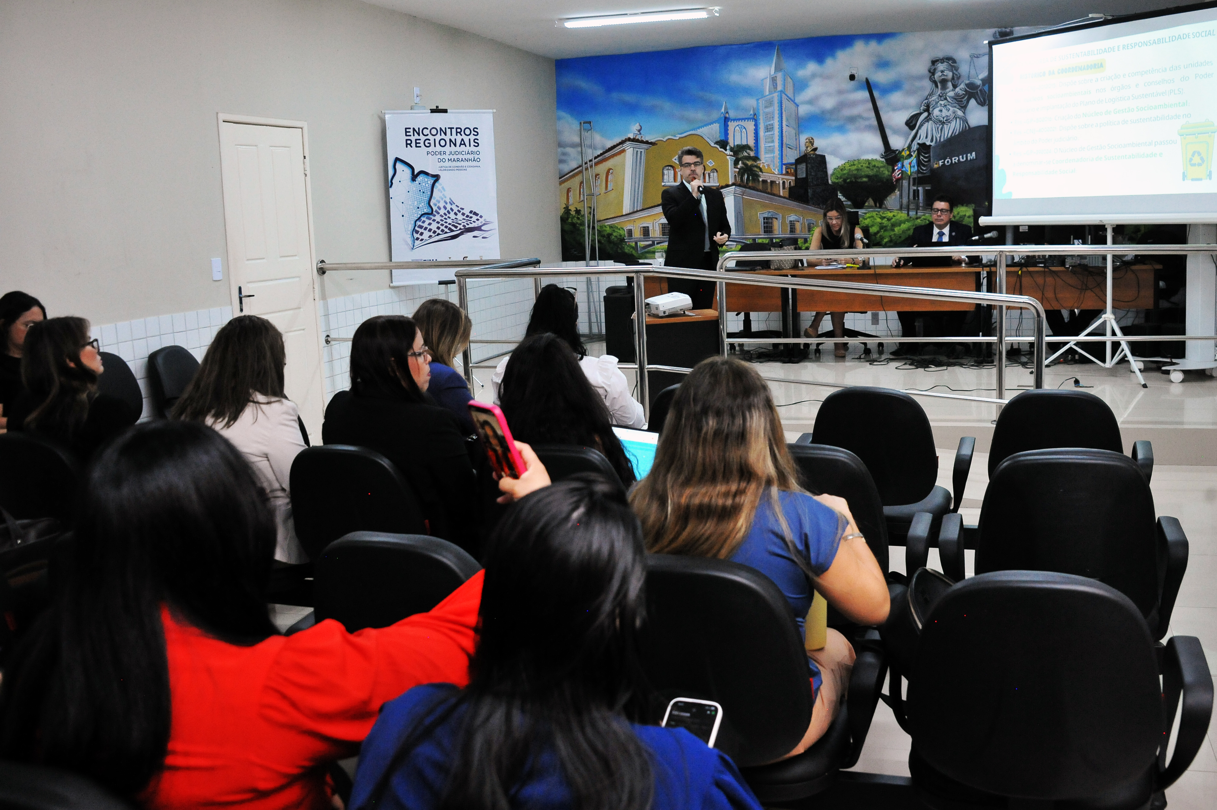 ## ¿ Descrição da Imagem: Evento em Sala de Aula/Auditório A imagem mostra um evento ou palestra acontecendo em um ambiente que se assemelha a uma **sala de aula ou pequeno auditório**. **1. O Palco/Mesa de Honra:** * No fundo da sala, há uma **mesa de honra** onde quatro pessoas estão sentadas. * Uma pessoa, vestindo um terno escuro, está de pé no centro, atrás da mesa, falando ao público. Ele parece ser o **palestrante** principal ou mediador no momento. * À sua direita, estão sentadas três outras pessoas (duas mulheres e um homem, todos com vestimentas formais ou semiformais). * Atrás da mesa, na parede, há um grande **mural** pintado com a imagem de uma paisagem urbana histórica (prédios, estátuas e o céu). * À direita do palestrante, um **projetor** exibe slides em uma tela branca. O texto na tela está em português e parece tratar de temas jurídicos ou sociais. **2. A Audiência:** * A maior parte da foto é preenchida pelo **público**, majoritariamente composto por mulheres, sentadas em cadeiras pretas de plástico/couro, dispostas em fileiras. * O público está atento ao palestrante. * Uma mulher em primeiro plano, vestindo uma blusa vermelha brilhante, e outra vestindo azul, estão visíveis de costas. * Algumas pessoas estão segurando seus celulares ou anotações. **3. Outros Elementos:** * Há um **banner/cartaz** pendurado à esquerda, que menciona "**ENCONTROS REGIONAIS**" e apresenta um desenho abstrato. * Uma porta branca está visível na parede lateral esquerda. Em resumo, a imagem captura um **momento formal de apresentação e debate**, com um palestrante ativo e um público engajado, ambientado por uma decoração que inclui um mural artístico e projeção de slides. --- Gostaria que eu tentasse identificar mais informações sobre o evento ou o local, pesquisando pelo texto visível nos cartazes, ou sobre o tema da palestra?