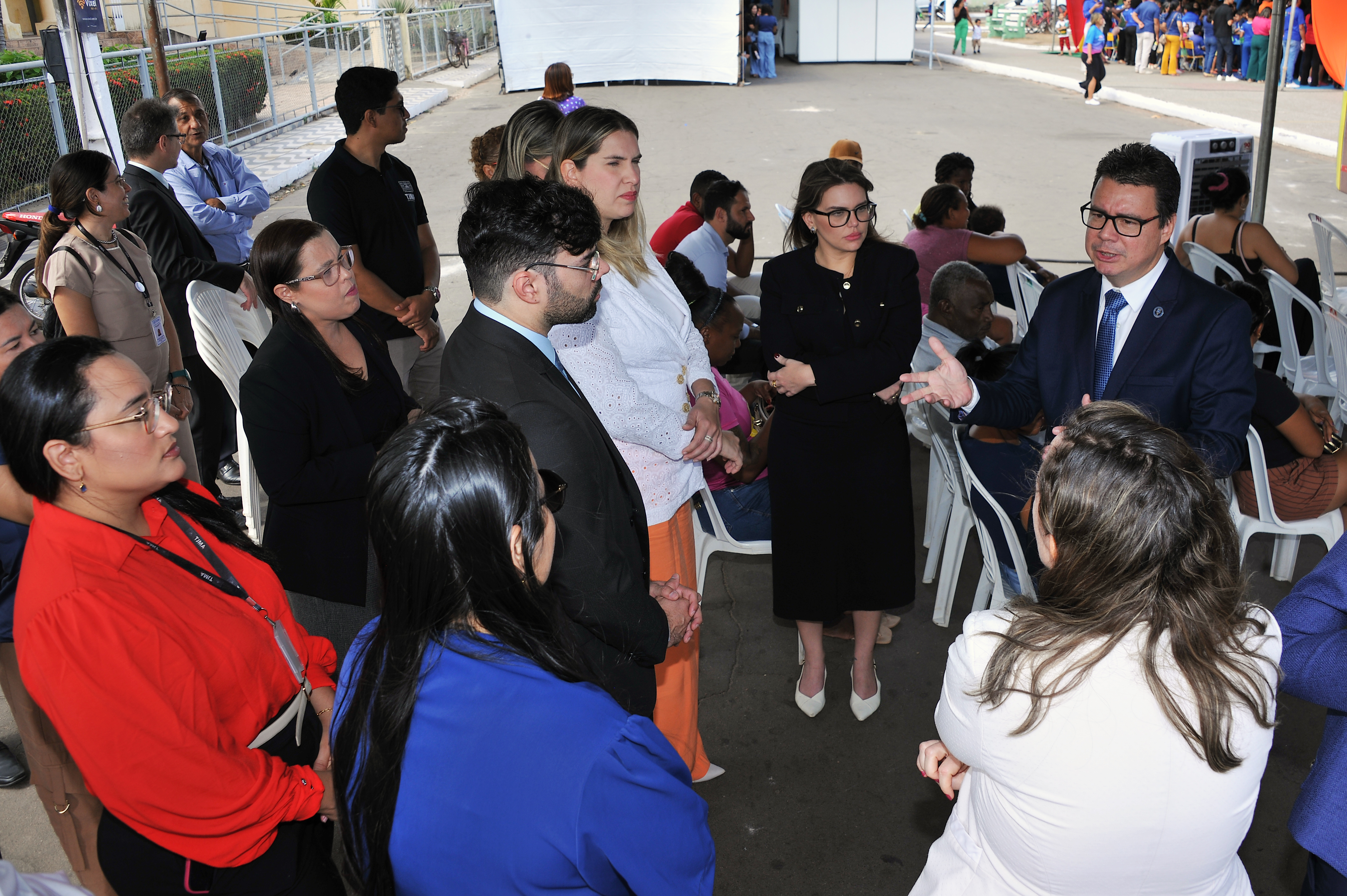 Esta imagem retrata uma **reunião ou discussão em grupo ao ar livre**, possivelmente em um evento, feira ou área de recepção. O foco principal está em um pequeno grupo de pessoas engajadas em uma conversa. ### ¿ Descrição Detalhada do Grupo Principal * **Homem de Terno (Orador/Líder):** Ele está em pé no lado direito, vestindo um terno escuro (azul marinho ou preto) com uma gravata azul e óculos. Ele gesticula com a mão direita, indicando que está explicando ou apresentando algo para o grupo. * **Mulher de Vestido Preto:** Em pé ao lado do homem de terno, ela veste um elegante vestido preto, tem óculos e sapatos de salto brancos. Ela está com os braços cruzados e parece estar escutando atentamente ou esperando sua vez de falar. * **Participantes Sentados e em Pé:** Várias outras pessoas, vestidas em trajes formais ou *business casual* (incluindo uma mulher de blusa vermelha vibrante à esquerda, e um homem de paletó escuro e óculos no centro), estão ao redor, olhando para o homem que fala. * Muitos estão sentados em cadeiras brancas de plástico. * A expressão geral do grupo é de **concentração e engajamento**. ### ¿ Descrição do Contexto e Fundo * **Ambiente:** A cena se passa em uma área externa pavimentada, possivelmente sob o sol, mas o grupo principal está em uma área de sombra parcial. * **Estruturas de Evento:** No fundo, podem ser vistas grandes estruturas de tendas brancas (ou *stands*) e grades metálicas, típicas de eventos ao ar livre. * **Público Distante:** Mais ao fundo, à direita, há um agrupamento maior de pessoas em roupas casuais e coloridas, sugerindo que o evento é maior e que esse grupo em primeiro plano pode ser de organizadores, autoridades, ou convidados especiais. * **Cercado/Fundo:** Há uma cerca metálica (grade) e algumas árvores e vegetação visíveis na parte superior esquerda. Em resumo, a imagem captura um momento de **comunicação formal ou de negócios** em um ambiente de evento público, onde um indivíduo está se dirigindo a um pequeno grupo de ouvintes atentos. Gostaria de ver outras imagens relacionadas a eventos ou reuniões?