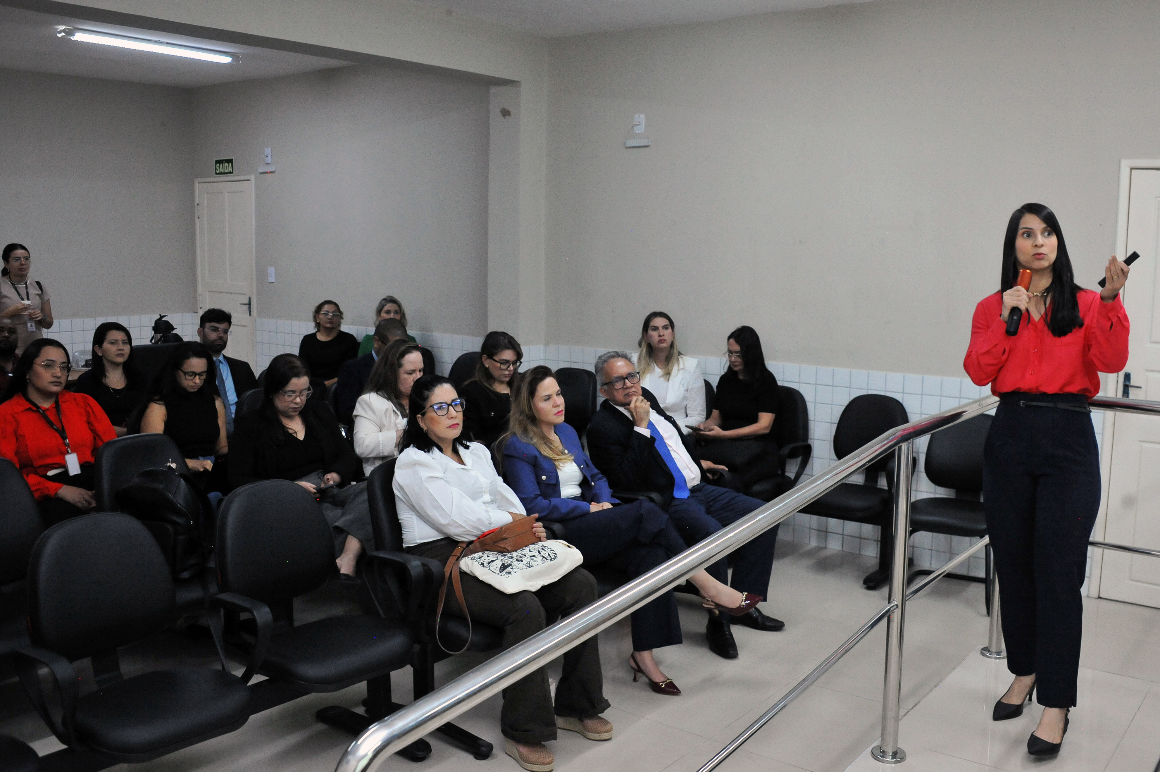 ## ¿ Descrição da Imagem: Palestra em Sala de Auditório A imagem retrata um **momento de apresentação ou palestra** em um ambiente interno que se assemelha a uma sala de reunião, auditório pequeno ou sala de treinamento. **1. A Palestrante:** * Uma mulher está de pé no lado direito da imagem, em posição de fala. * Ela veste uma **camisa social vermelha** vibrante e calças sociais escuras (pretas ou azul-marinho). * Ela tem cabelo escuro e longo e segura um microfone (vermelho) na mão esquerda e um objeto de controle remoto/apontador na mão direita. * Sua postura é de quem está **expondo um conteúdo** para a plateia. **2. A Audiência:** * O público está sentado em cadeiras pretas (semelhantes às da imagem anterior) dispostas em fileiras no lado esquerdo e no centro. * A audiência é composta por homens e mulheres, a maioria vestindo roupas escuras (preto ou cores sóbrias) e formais/semiformais, como é comum em eventos corporativos ou jurídicos. * O foco do público está voltado para a palestrante. * Há uma **grade metálica** de proteção ou apoio que separa levemente a área da palestrante da plateia. **3. O Ambiente:** * As paredes são de cor clara (bege ou cinza claro), e o teto possui iluminação fluorescente. * Há um **revestimento em azulejo branco** na parte inferior da parede, atrás da plateia. * No fundo, à esquerda, há uma **porta branca** com um sinal de "SAÍDA" verde acima dela. * O ambiente sugere um espaço **institucional ou profissional**, com decoração simples e funcional. Em resumo, a foto captura a **interação entre uma palestrante energizada e um público atento**, em um ambiente profissional e bem iluminado. --- Posso procurar informações sobre a organização que utiliza esse tipo de espaço ou sobre o possível tema da palestra (se houver alguma pista no contexto das imagens) se desejar.