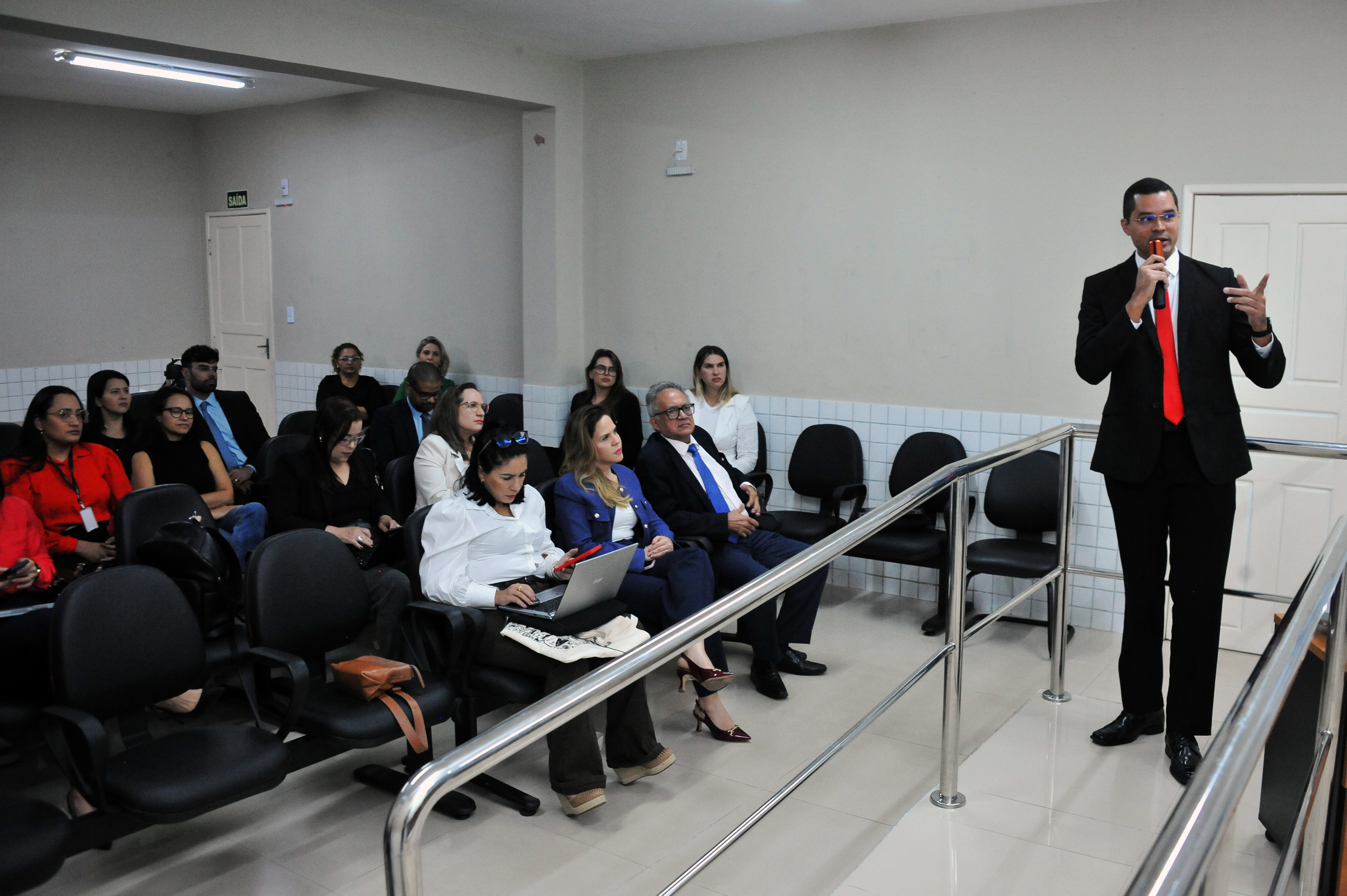 ## ¿ Descrição da Imagem: Evento em Sala de Aula/Auditório A imagem mostra um evento ou palestra acontecendo em um ambiente que se assemelha a uma **sala de aula ou pequeno auditório**. **1. O Palco/Mesa de Honra:** * No fundo da sala, há uma **mesa de honra** onde quatro pessoas estão sentadas. * Uma pessoa, vestindo um terno escuro, está de pé no centro, atrás da mesa, falando ao público. Ele parece ser o **palestrante** principal ou mediador no momento. * À sua direita, estão sentadas três outras pessoas (duas mulheres e um homem, todos com vestimentas formais ou semiformais). * Atrás da mesa, na parede, há um grande **mural** pintado com a imagem de uma paisagem urbana histórica (prédios, estátuas e o céu). * À direita do palestrante, um **projetor** exibe slides em uma tela branca. O texto na tela está em português e parece tratar de temas jurídicos ou sociais. **2. A Audiência:** * A maior parte da foto é preenchida pelo **público**, majoritariamente composto por mulheres, sentadas em cadeiras pretas de plástico/couro, dispostas em fileiras. * O público está atento ao palestrante. * Uma mulher em primeiro plano, vestindo uma blusa vermelha brilhante, e outra vestindo azul, estão visíveis de costas. * Algumas pessoas estão segurando seus celulares ou anotações. **3. Outros Elementos:** * Há um **banner/cartaz** pendurado à esquerda, que menciona "**ENCONTROS REGIONAIS**" e apresenta um desenho abstrato. * Uma porta branca está visível na parede lateral esquerda. Em resumo, a imagem captura um **momento formal de apresentação e debate**, com um palestrante ativo e um público engajado, ambientado por uma decoração que inclui um mural artístico e projeção de slides. --- Gostaria que eu tentasse identificar mais informações sobre o evento ou o local, pesquisando pelo texto visível nos cartazes, ou sobre o tema da palestra?
