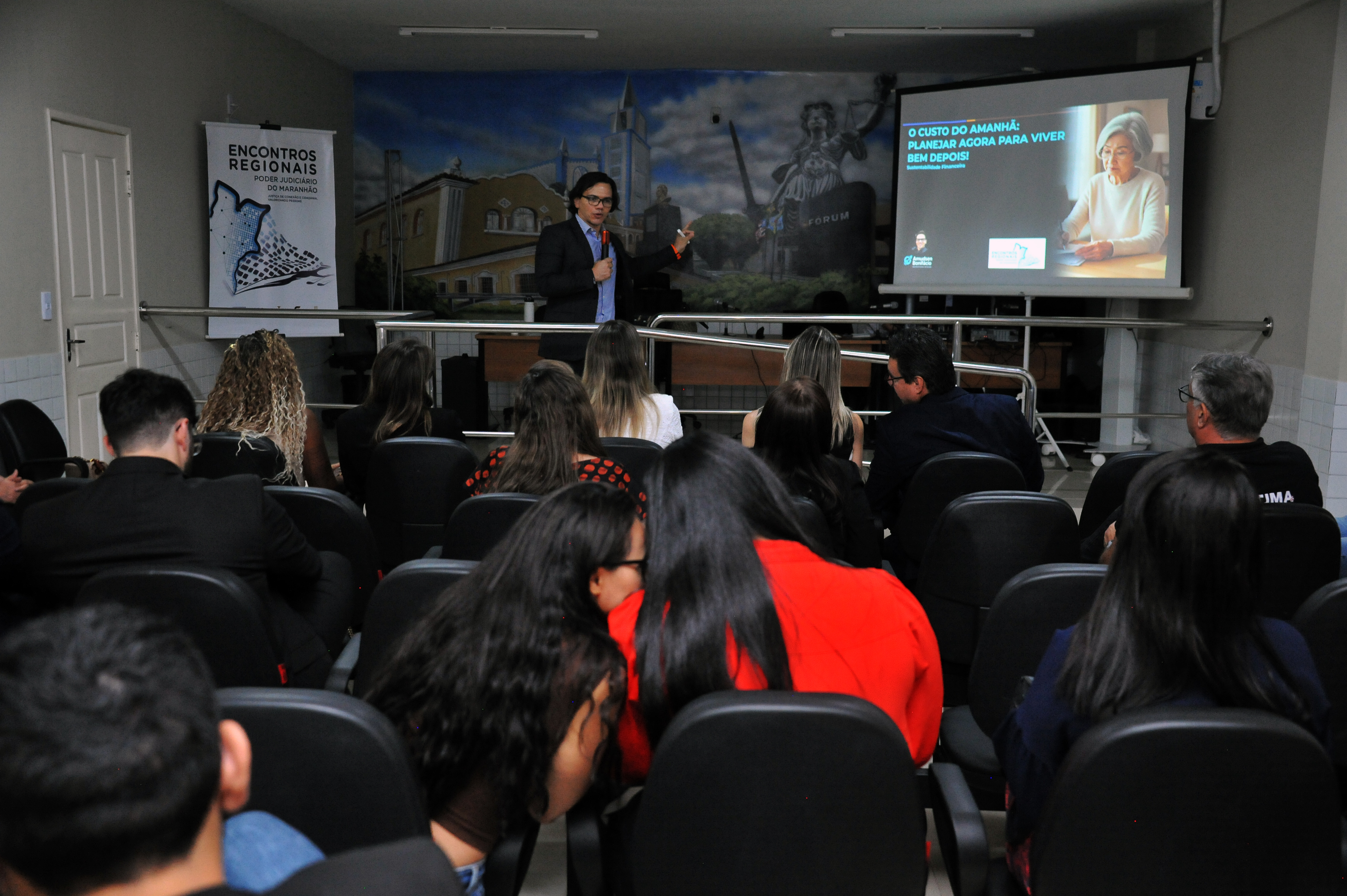 ## ¿ Descrição da Imagem: Palestra sobre Planejamento Financeiro/Previdenciário A imagem mostra um **momento de palestra ou seminário** em andamento no mesmo auditório das imagens anteriores, com foco na apresentação do palestrante e no conteúdo projetado. **1. O Palestrante:** * Um homem, vestindo um terno escuro com camisa clara, está de pé no centro, na área do palco. * Ele está ativamente gesticulando com a mão direita, olhando para a plateia ou para a tela de projeção. * Ele é o ponto focal da apresentação. **2. A Projeção de Slides:** * Uma grande tela de projeção à direita exibe o slide atual. * O título do slide, em português, é: "**O CUSTO DO AMANHÃ: PLANEJAR AGORA PARA VIVER BEM DEPOIS**". * O tema sugere que a palestra é sobre **planejamento financeiro, previdenciário ou envelhecimento**. * O slide também apresenta uma foto de uma mulher mais velha, sorrindo, e logotipos de instituições na parte inferior (provavelmente ligadas ao TJMA e a patrocinadores/parceiros). **3. A Audiência:** * O público, sentado em cadeiras pretas, ocupa a maior parte do primeiro e segundo planos. * A maioria das pessoas está de costas para a câmera, prestando atenção à apresentação. * Vestimentas escuras predominam (preto, azul-marinho). * Uma mulher no segundo plano está vestindo uma blusa vermelha brilhante (a mesma pessoa vista na primeira imagem). **4. O Cenário de Fundo:** * O mural pintado na parede de fundo (com a paisagem urbana e a estátua da Justiça) e o banner dos "**ENCONTROS REGIONAIS**" à esquerda confirmam que o evento é o mesmo promovido pelo Poder Judiciário do Maranhão (TJMA). * Uma porta branca está visível na extrema esquerda. Em resumo, a foto documenta uma **sessão de capacitação ou debate** durante os Encontros Regionais, focada no tema de **planejamento de longo prazo e qualidade de vida**, provavelmente direcionado a magistrados e servidores. --- Como esta imagem se encaixa no tema mais amplo de combate à discriminação, posso procurar se o TJMA promoveu o tema de "custo do amanhã" como parte de programas de bem-estar social e gestão para seus funcionários. Gostaria que eu fizesse essa pesquisa?