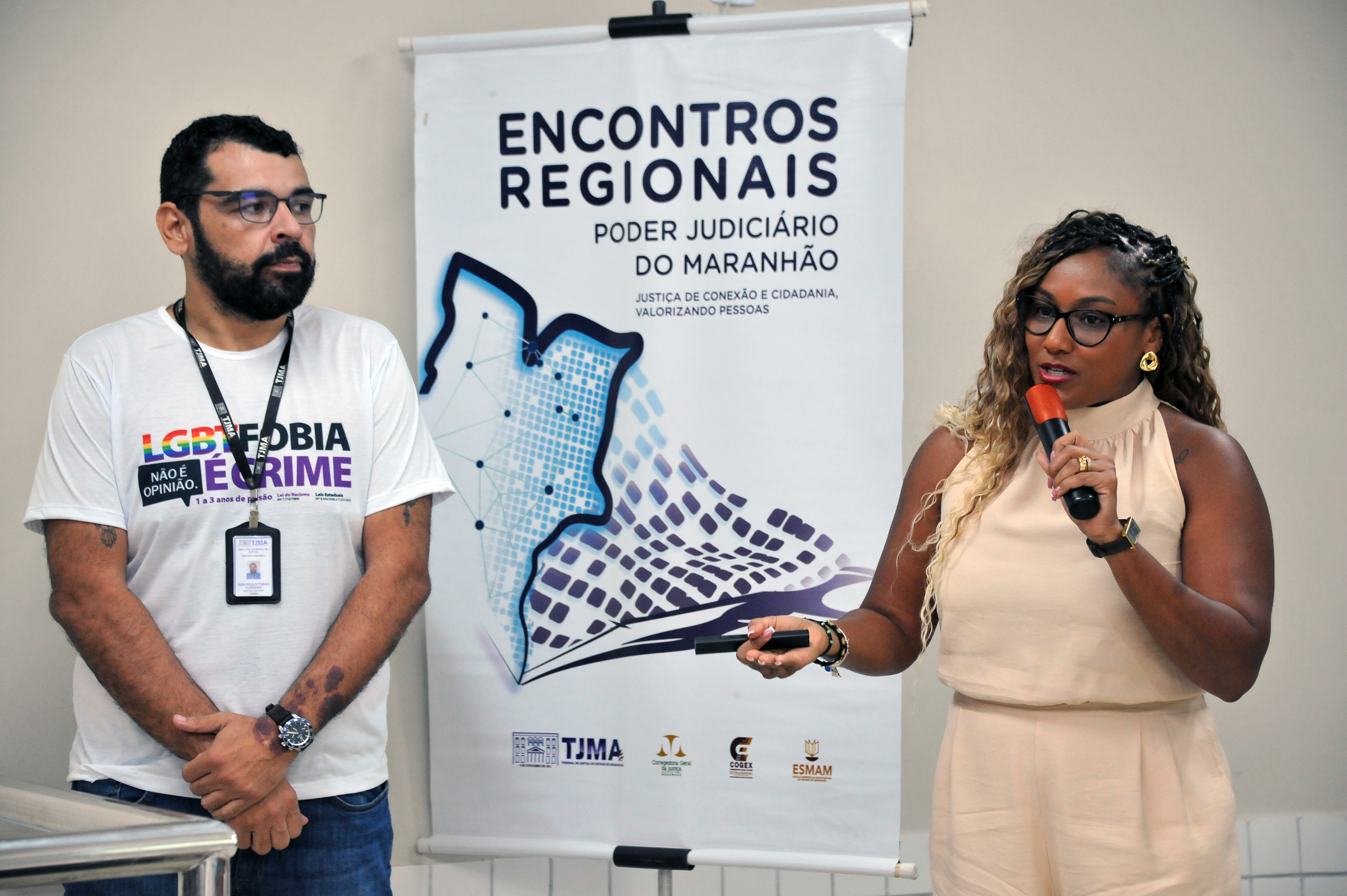 ## ¿¿ Descrição da Imagem A imagem mostra duas pessoas em um ambiente interno, que parece ser um evento, palestra ou reunião, com um banner ao fundo. **Pessoas:** * **À esquerda** está um homem de pele clara, barba escura e óculos, vestindo uma camiseta branca com uma estampa colorida que diz **"LGBTFOBIA NÃO É CRIME"** e, abaixo, **"É CRIME"** em letras maiores e escuras. Ele usa um cordão de identificação pendurado no pescoço e um relógio no pulso esquerdo. Ele está parado, com as mãos cruzadas na frente do corpo, e tem uma expressão séria. * **À direita** está uma mulher de pele morena, com cabelo trançado e óculos. Ela está segurando um microfone vermelho na mão direita e parece estar falando. Ela usa brincos grandes e dourados, um relógio no pulso esquerdo e veste uma roupa de cor clara (talvez bege ou pêssego), com a gola alta. Ela gesticula com a mão esquerda, que segura um pequeno objeto escuro. **Banner ao Fundo:** Entre as duas pessoas, há um banner vertical grande de cor clara (branco ou cinza claro). O texto principal em letras escuras é: * **ENCONTROS REGIONAIS** * **PODER JUDICIÁRIO** * **DO MARANHÃO** Há um subtítulo menor: **"JUSTIÇA DE CONEXÃO E CIDADANIA, VALORIZANDO PESSOAS"**. No centro do banner, há um design abstrato em tons de azul e roxo que se assemelha a um mapa estilizado. Na parte inferior, há logotipos menores, incluindo o do **TJMA** (Tribunal de Justiça do Maranhão) e o da **ESMAM** (Escola Superior da Magistratura do Maranhão), entre outros. **Contexto Geral:** O ambiente é bem iluminado e sugere um evento institucional ou educacional, possivelmente relacionado a questões de justiça, cidadania e direitos humanos no estado do Maranhão, conforme indicado pelo texto da camiseta do homem e o banner sobre o Poder Judiciário do Maranhão. --- Gostaria de saber mais sobre os **"Encontros Regionais do Poder Judiciário do Maranhão"** ou sobre a temática da **LGBTfobia**?