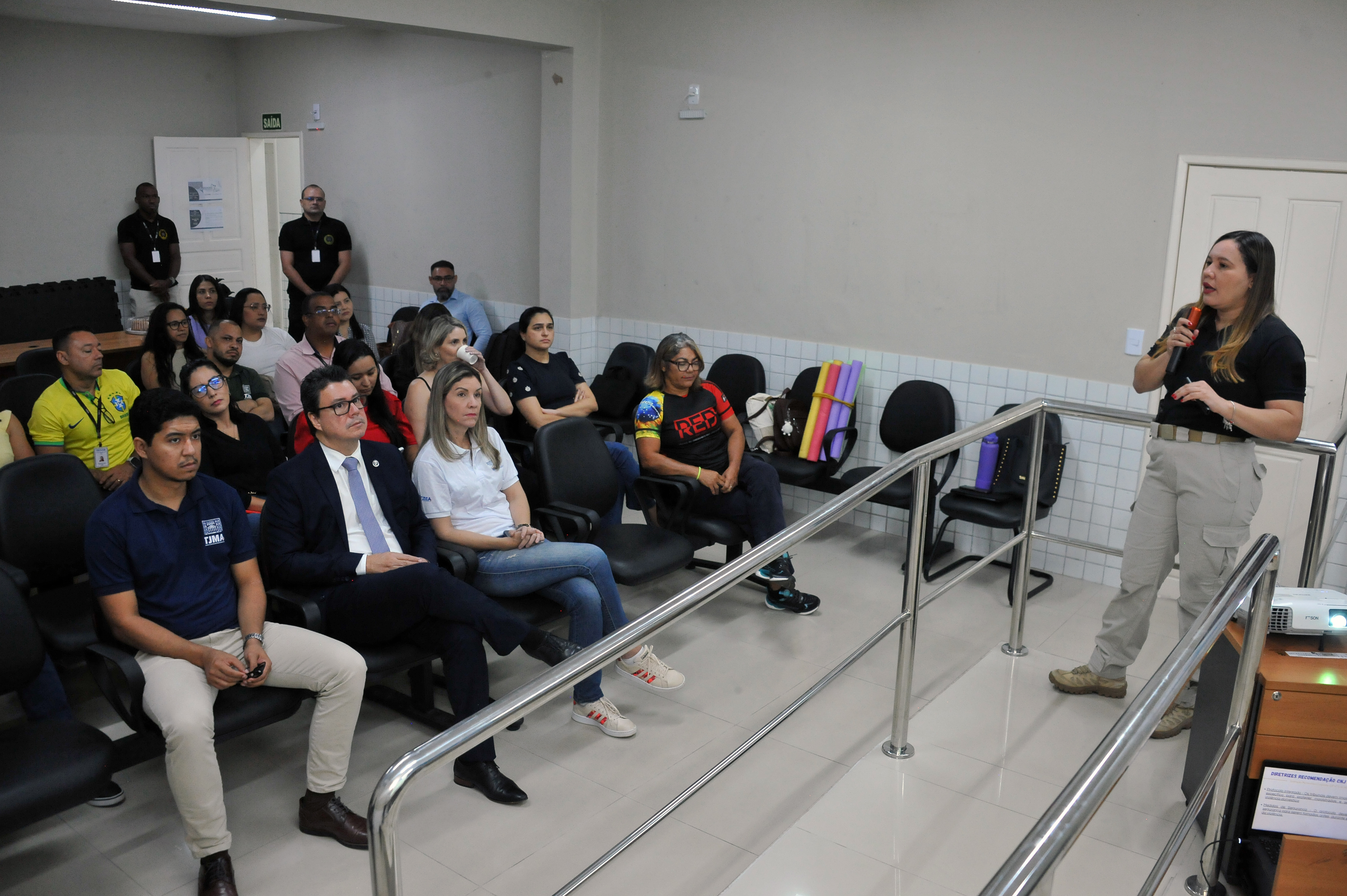 Essa imagem parece ser de um **evento, palestra, ou reunião** em um ambiente interno, possivelmente um tribunal, sala de audiências ou sala de conferências.  Aqui está uma descrição detalhada:  * **Pessoa Falando:** No lado direito da imagem, uma mulher de cabelos loiros/grisalhos e óculos está em pé, segurando um microfone na mão direita e gesticulando com a esquerda, parecendo estar **apresentando ou discursando** para o grupo sentado. Ela veste uma camisa preta e calças cáqui (ou bege) de estilo utilitário e botas. * **Público:** Um grupo de **aproximadamente 20 pessoas** está sentado em cadeiras de plástico pretas, voltado para a oradora.     * As primeiras filas são compostas por algumas pessoas vestindo trajes sociais (um homem de terno escuro) e casuais (homens e mulheres com camisas polo, camisas brancas e pretas, e jeans/calças claras).     * No canto inferior esquerdo, um homem de camisa polo azul-marinho e calças claras está sentado na primeira fila. * **Ambiente:** A sala tem paredes de cor clara/creme. Há um **corrimão metálico** (possivelmente de alumínio ou aço inoxidável) separando a oradora da primeira fila do público. No fundo, é possível ver uma porta com a palavra "SAÍDA" escrita acima dela. O ambiente é simples e funcional. * **Detalhes:** Há alguns objetos visíveis nas cadeiras vazias no fundo, como um tapete de ioga enrolado e um casaco/mochila. Outras pessoas, incluindo um homem de terno escuro, estão visíveis no canto superior esquerdo, perto da porta.  Em resumo, a foto captura um **momento de comunicação formal ou instrucional**.  Gostaria de saber mais detalhes sobre algum aspecto específico da foto?