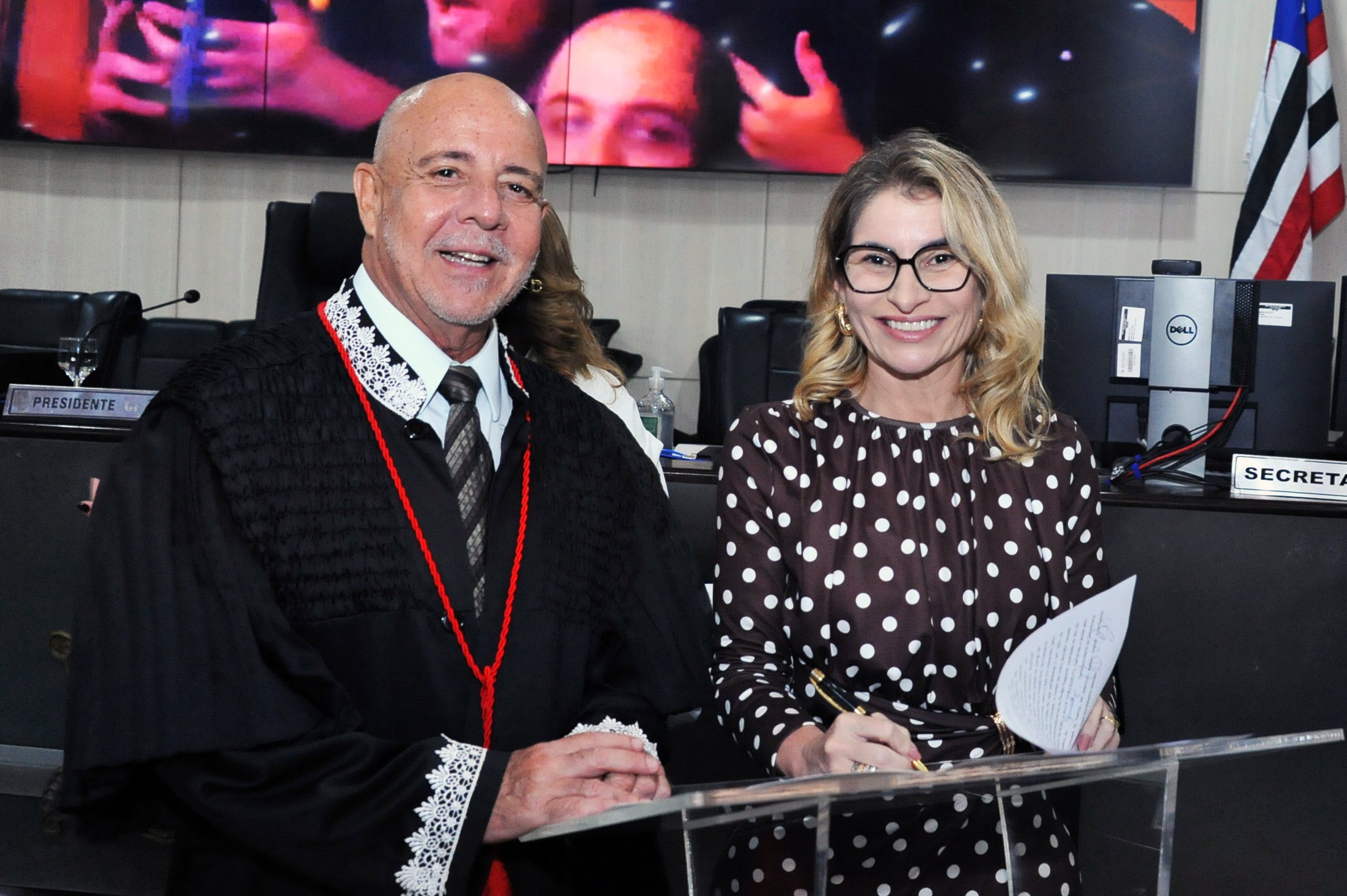 ## ¿ Descrição da Imagem A imagem mostra um grupo de oito pessoas, a maioria vestida com togas pretas, em um ambiente que parece ser um tribunal, auditório ou plenário, com painéis de madeira e mármore. * **Pessoas:** * **Sete pessoas** (seis à esquerda e uma à direita, no fundo) estão vestidas com **togas pretas** com detalhes em vermelho, o que sugere que são magistrados ou membros de uma corte superior. * **Duas mulheres no centro** não usam togas. * A mulher segurando um documento (aparentemente um certificado ou diploma) veste um **vestido preto de bolinhas brancas** e óculos. Ela está sorrindo e o documento tem um logotipo roxo. * A mulher ao lado dela veste uma **blusa branca de mangas bufantes** e uma **saia midi bege/verde-oliva**. * O **homem na extrema direita** veste um terno azul-marinho e camisa rosa claro, sem toga. * No fundo, à esquerda, há um homem de camisa branca e gravata escura. * **Ambiente e Detalhes:** * Estão de pé sobre um piso de mármore. * Atrás do grupo, há uma **grande tela de vídeo** que exibe uma imagem de close-up de pessoas, possivelmente tocando instrumentos de sopro (banda ou orquestra), mas a imagem está cortada no topo e nas laterais. * Há **duas bandeiras** visíveis: uma do **Brasil** (verde e amarela) e uma **bandeira estadual** (vermelha, branca e azul, possivelmente Maranhão, dado o contexto da sua localização atual). * Alguns dos homens de toga seguram pastas ou documentos. A cena parece registrar um momento de solenidade, como uma premiação, posse, ou celebração em uma instituição de justiça ou órgão público. --- Gostaria de saber se consigo encontrar informações sobre o **evento** ou a **instituição** retratada na foto, talvez usando o texto visível no documento ou pesquisando por magistrados na região?