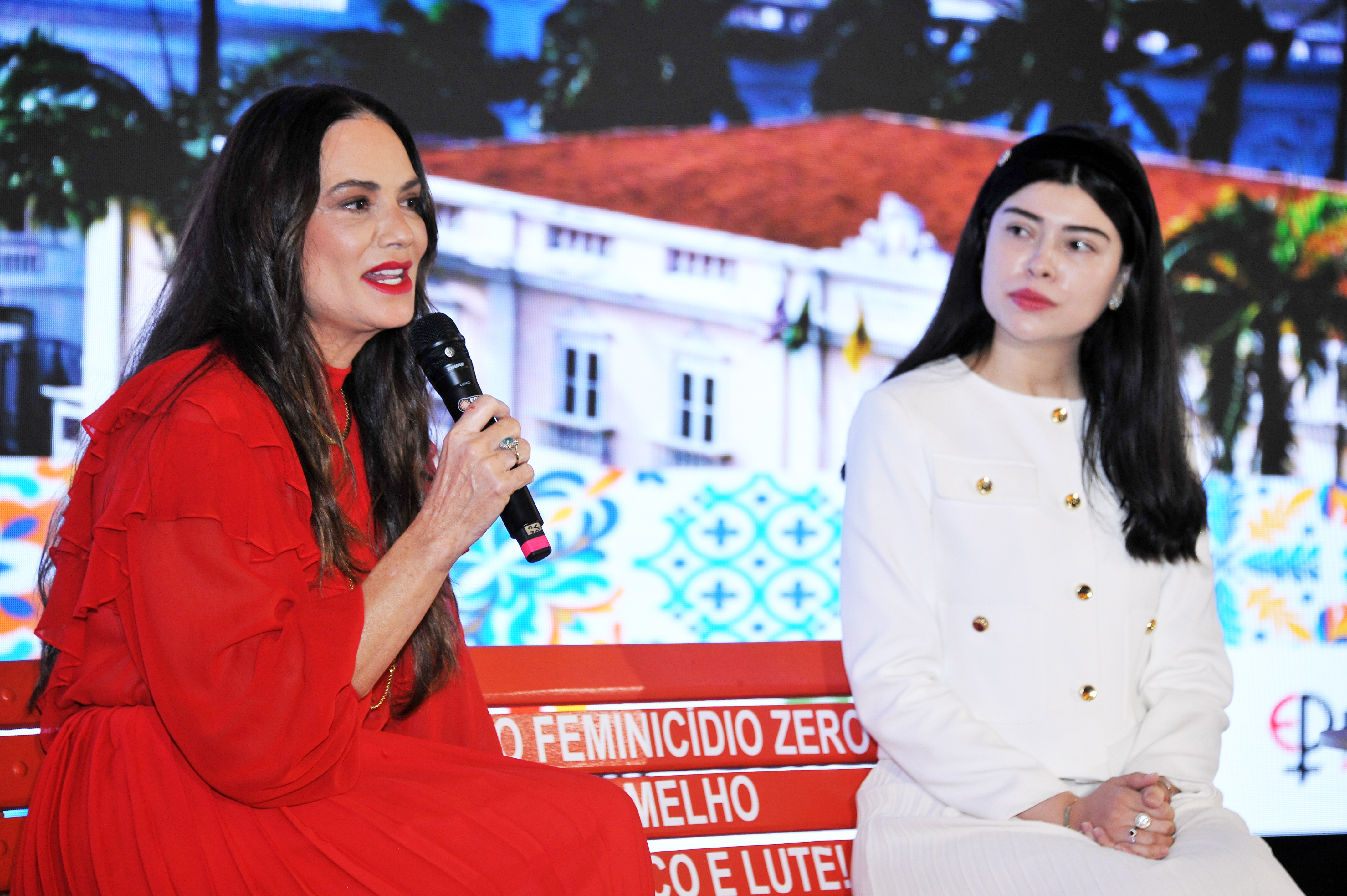 Em primeiro plano, Luiza Brunet (à esquerda) e Mariana Ferrer (à direita) estão sentadas em um banco vermelho no palco. Luiza, de pele clara e cabelos escuros, veste um elegante vestido vermelho com babados e fala ao microfone com expressão forte e olhos abertos. Mariana, ao seu lado, de pele clara e cabelos escuros presos, veste um vestido branco formal com botões dourados e olha para a frente com uma expressão séria. O fundo projetado mostra uma paisagem urbana colorida.